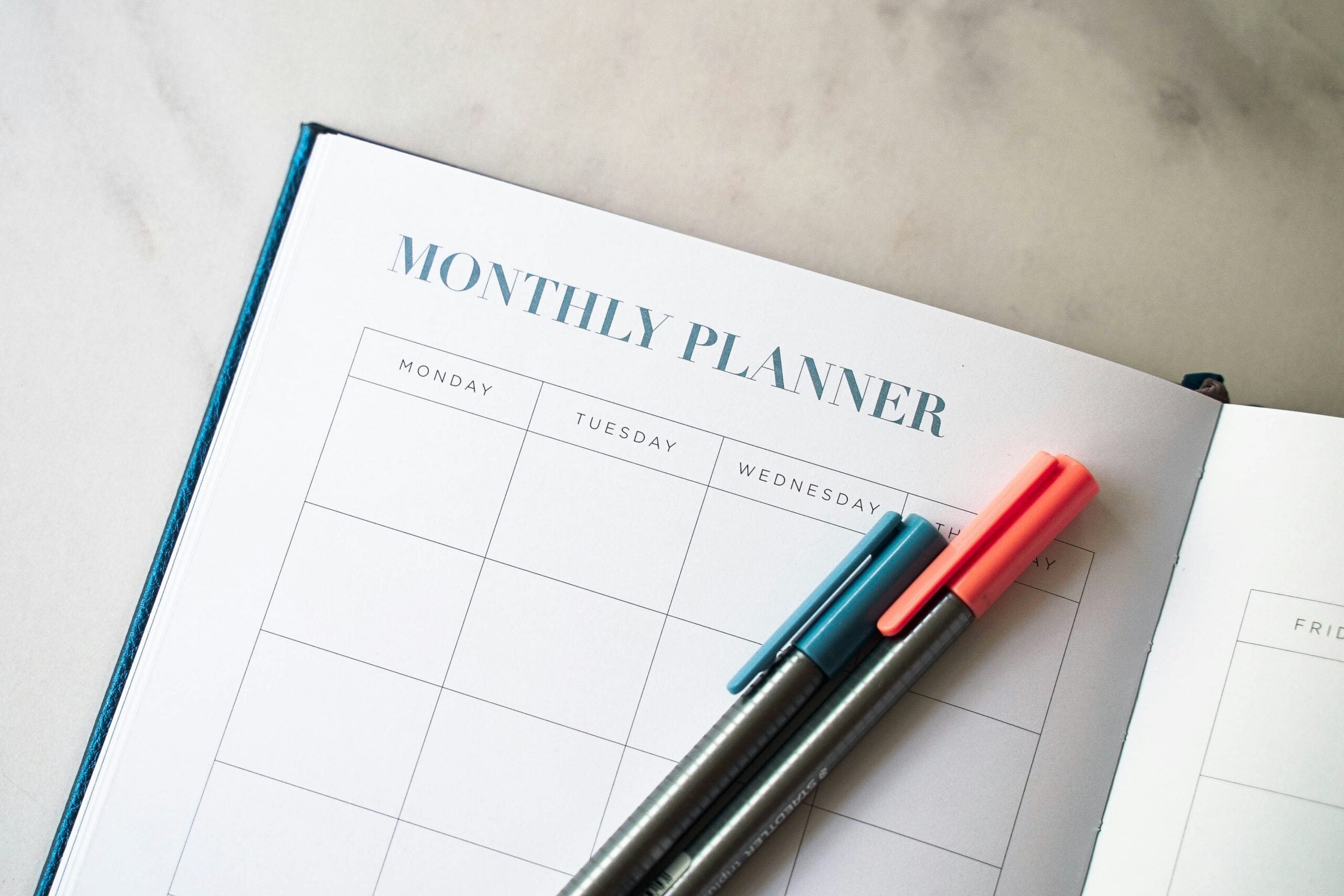 Open monthly planner showing a calendar grid for Monday to Friday. Two pens, one black and one coral, rest on the planner. The background is a light, smooth surface.