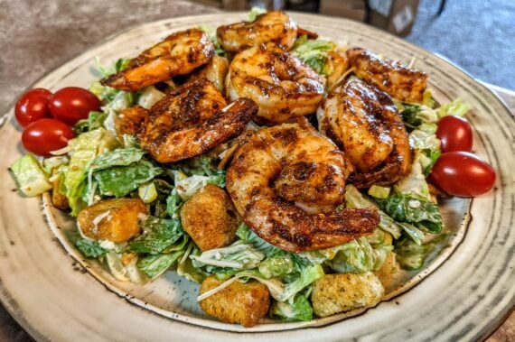 Grilled shrimp on a bed of Caesar salad with romaine lettuce, croutons, cherry tomatoes, and creamy dressing, served on a rustic plate.