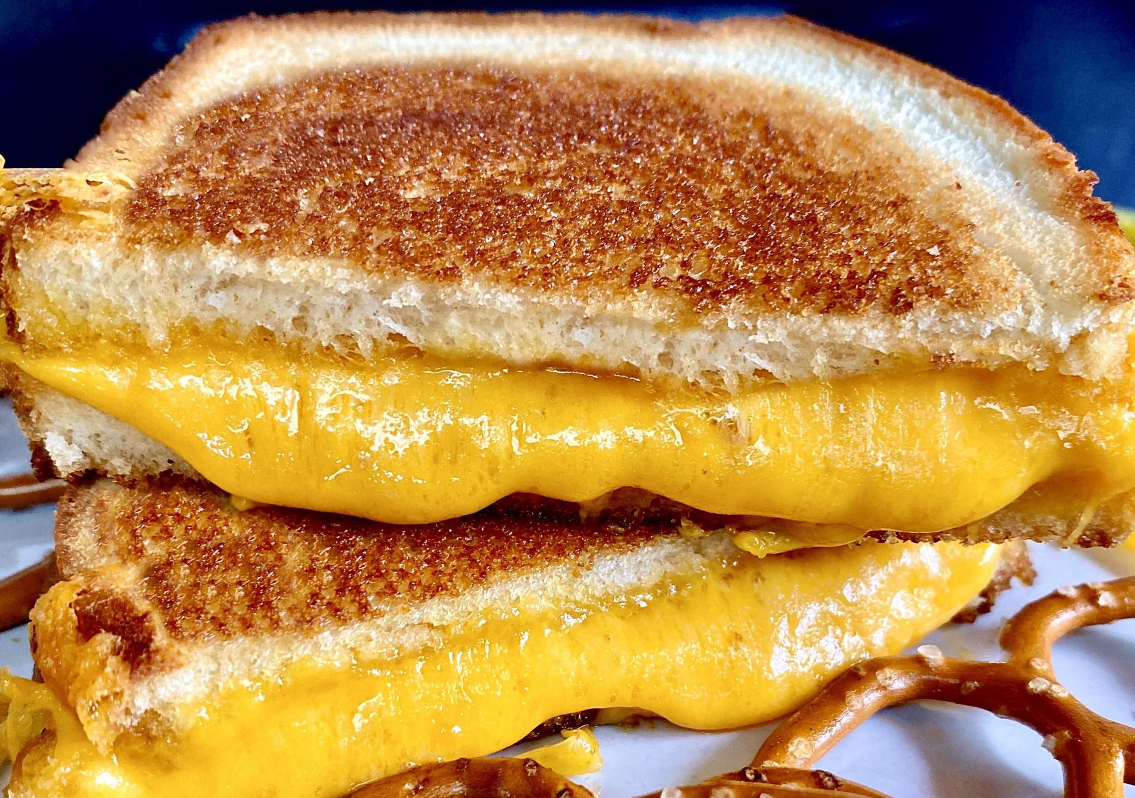 A close-up of Grilled Cheese Sandwiches with golden, toasted bread and melted cheddar cheese, stacked on a plate with pieces of pretzel underneath.