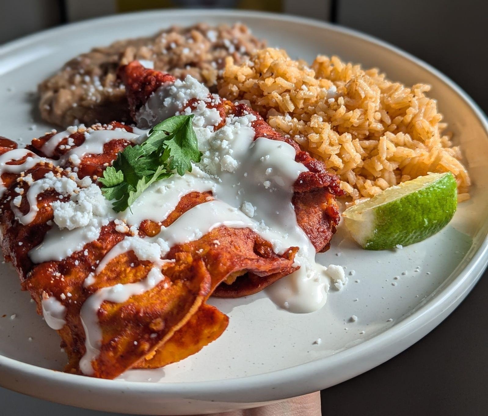 Three enchiladas topped with crema, cheese, and cilantro, served with Mexican rice, refried beans, and a lime wedge on a white plate.