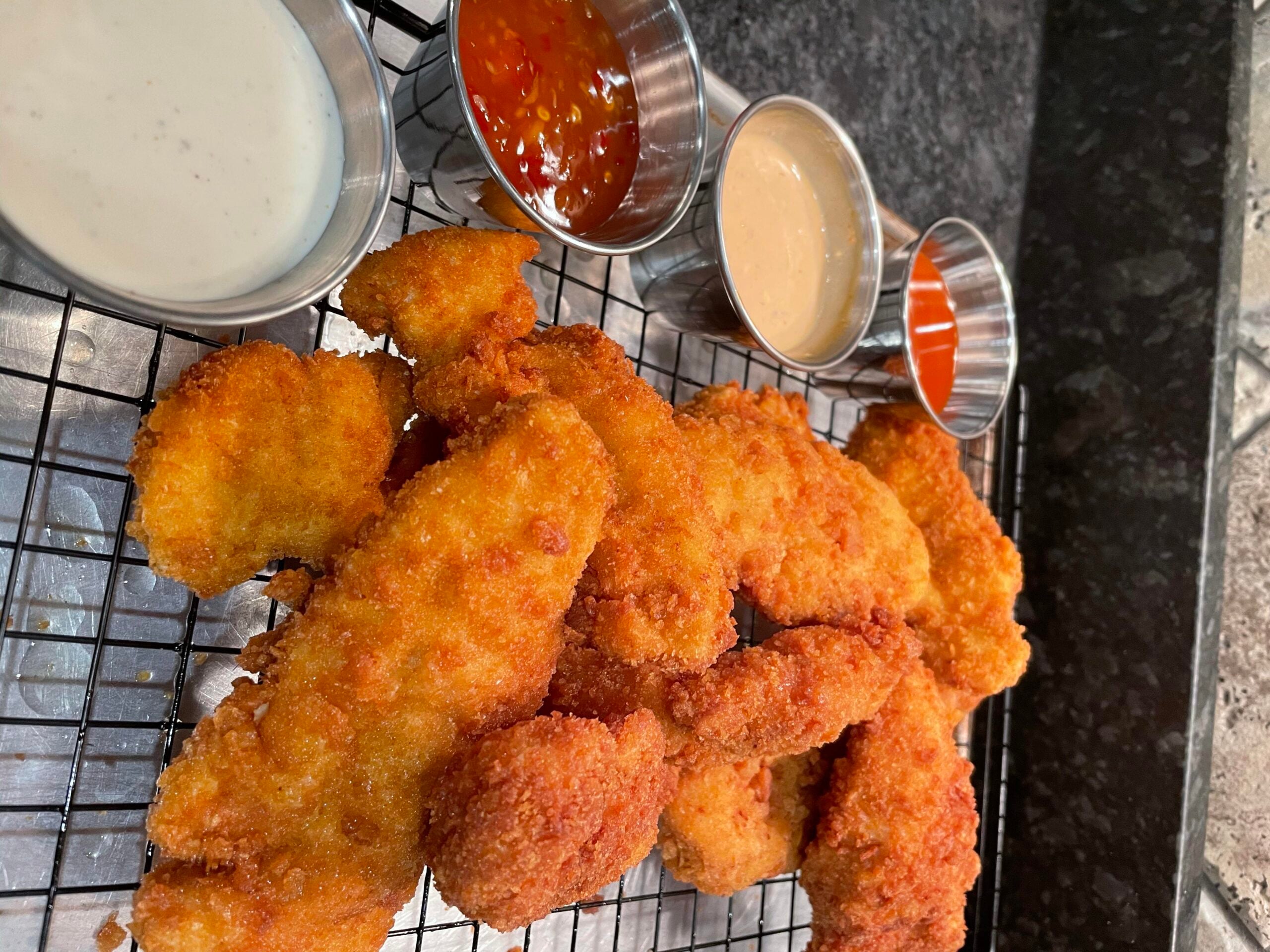 Golden fried chicken tenders on a cooling rack with four metal cups of dipping sauces, including ranch, sweet chili, honey mustard, and buffalo sauce, on a dark countertop.