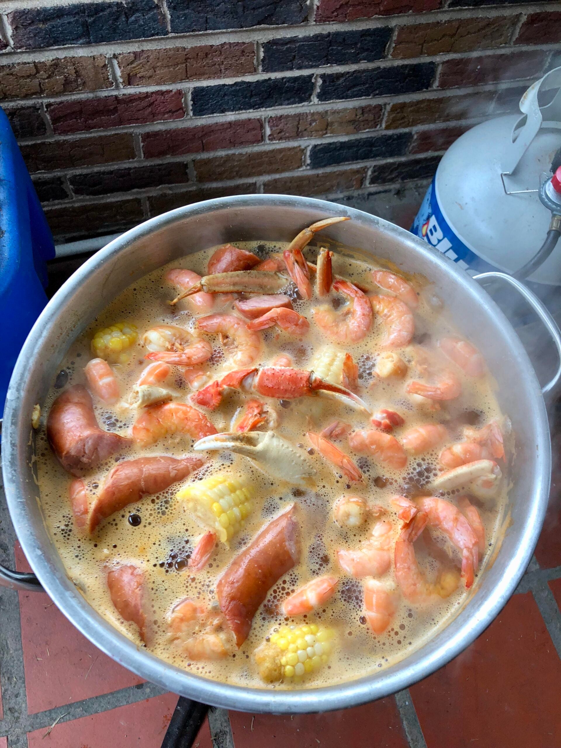 A large pot of seafood boil with shrimp, sausage, crab legs, and corn on the cob cooking outdoors, with steam rising and a propane tank nearby.