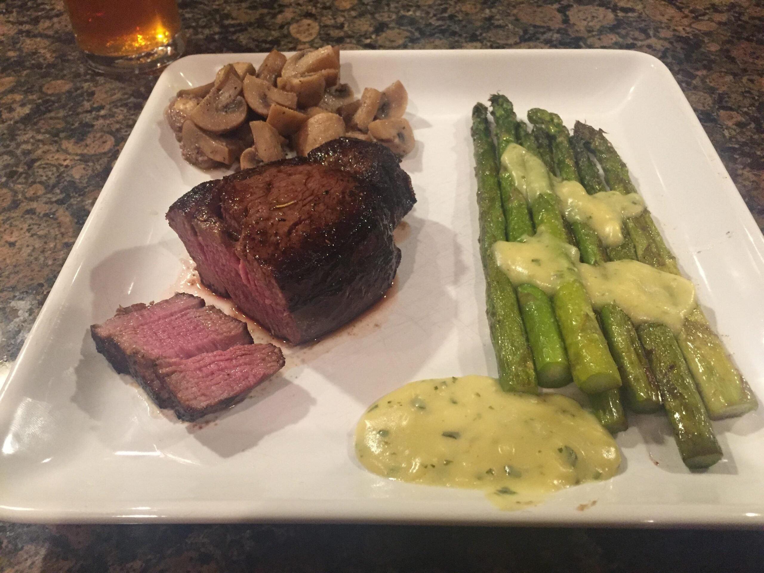 Grilled steak with a slice cut, sautéed mushrooms, and asparagus topped with a creamy sauce, served on a square white plate on a brown patterned countertop.