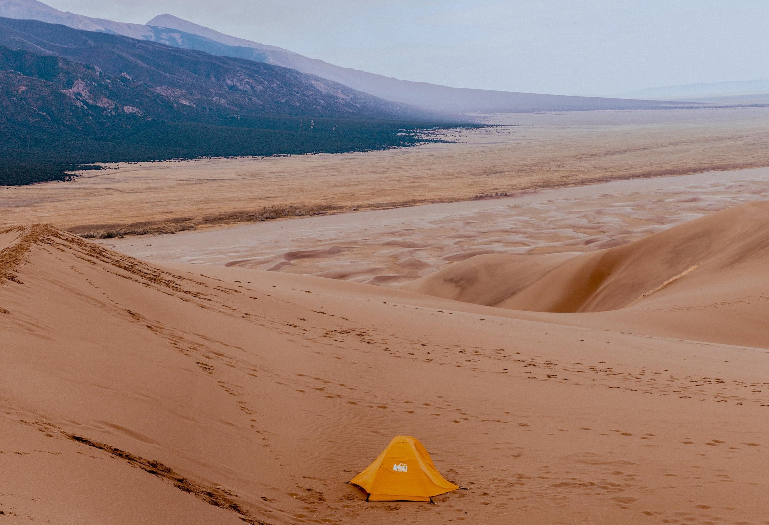 A single yellow tent sits on sand dunes with mountains and a vast valley in the background under a hazy sky.
