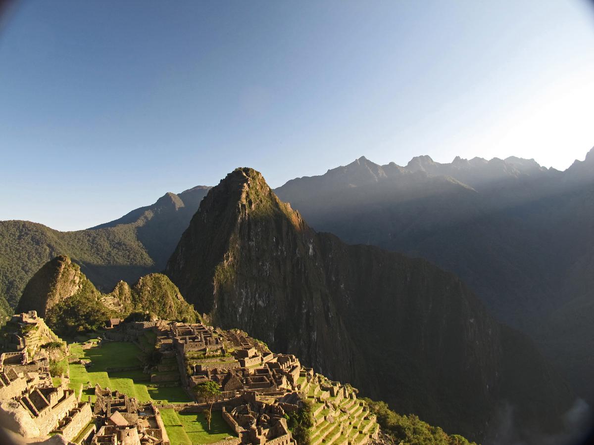 Machu Picchu’s ancient stone ruins sit on a mountain ridge under clear blue sky, surrounded by lush green peaks and dramatic shadows cast by the setting sun.
