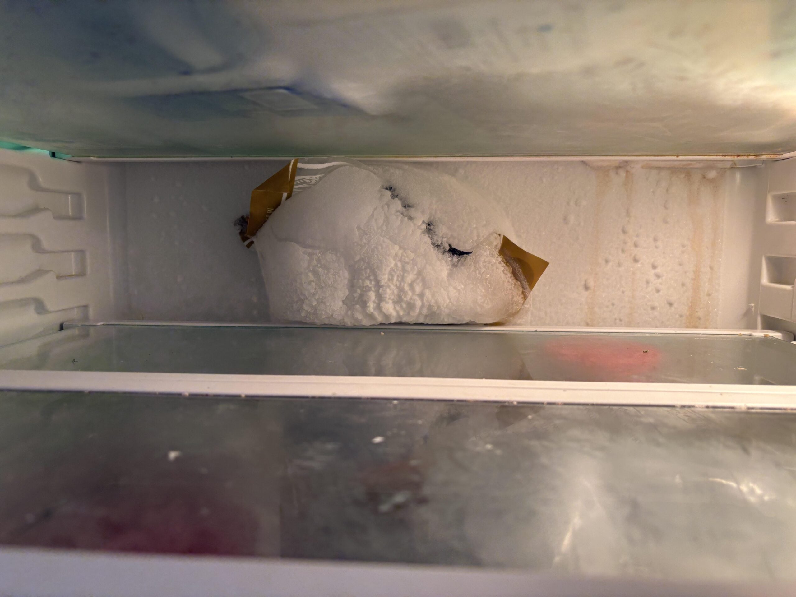 A frosted-over bag with yellow tape is stuck to the back wall of a freezer, surrounded by ice buildup. The freezer shelves are mostly empty, with some frost on the surfaces.