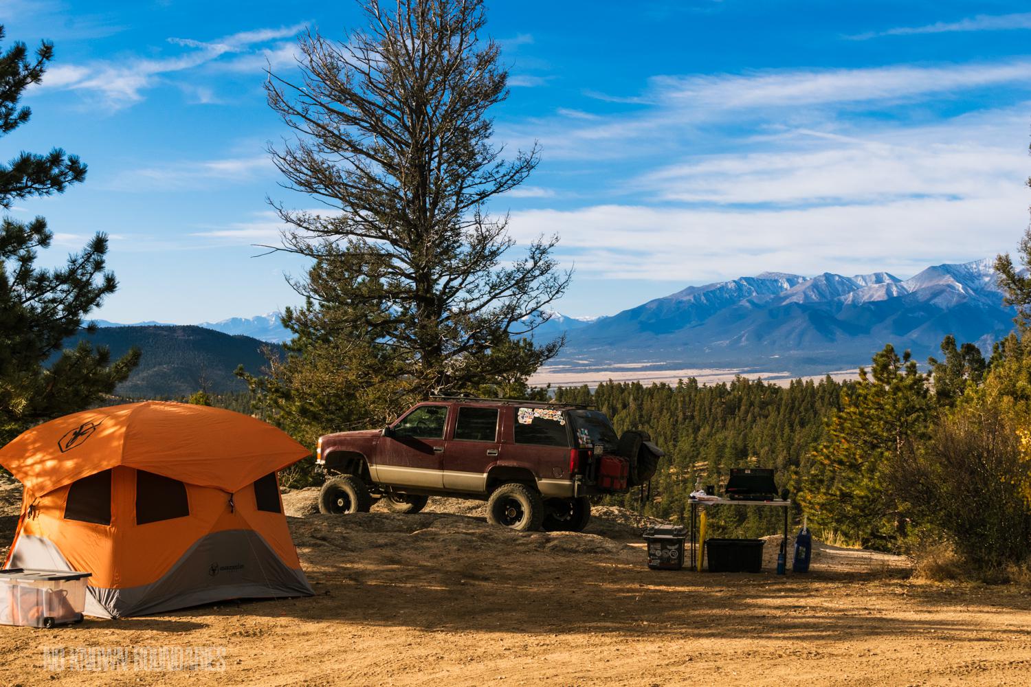 Orange tent and red SUV parked on a dirt clearing surrounded by pine trees, with camping gear nearby and snow-capped mountains visible in the distance under a blue sky.