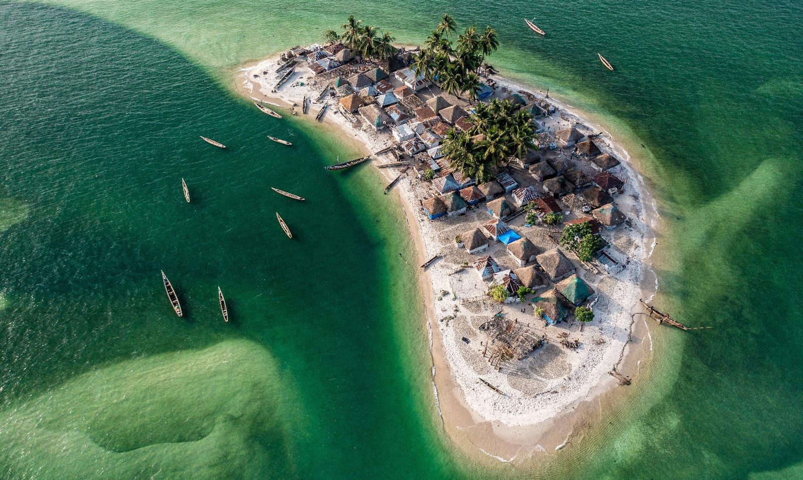 Aerial view of a small tropical island with clusters of thatched-roof huts, surrounded by green water and several wooden boats floating nearby, all set against the backdrop of incredible beaches.
