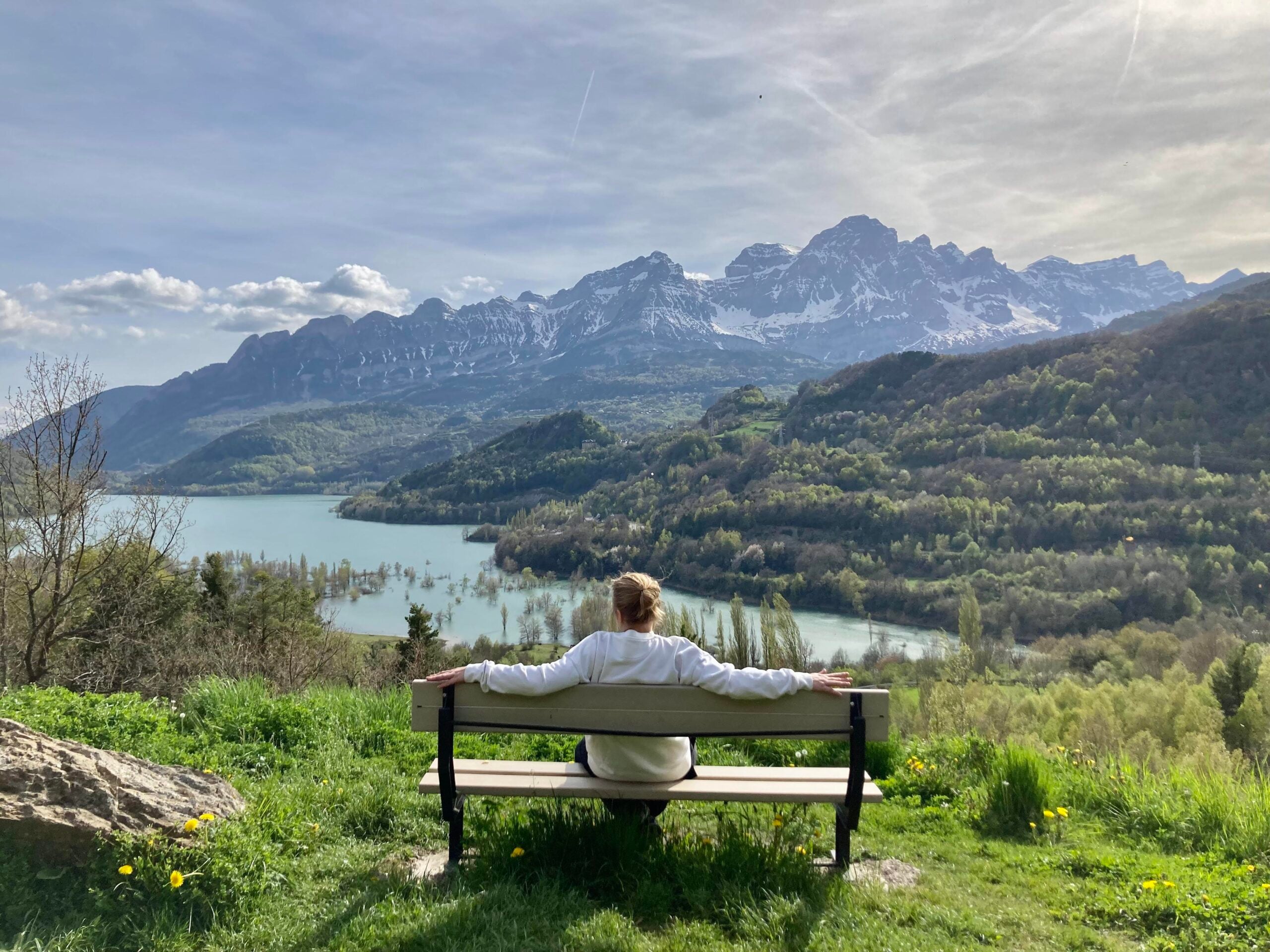 A person sits on a bench facing a scenic view of a lake, lush green hills, and snow-capped mountains under a cloudy sky—capturing the tranquil moment often seen in photos posted by hikers.