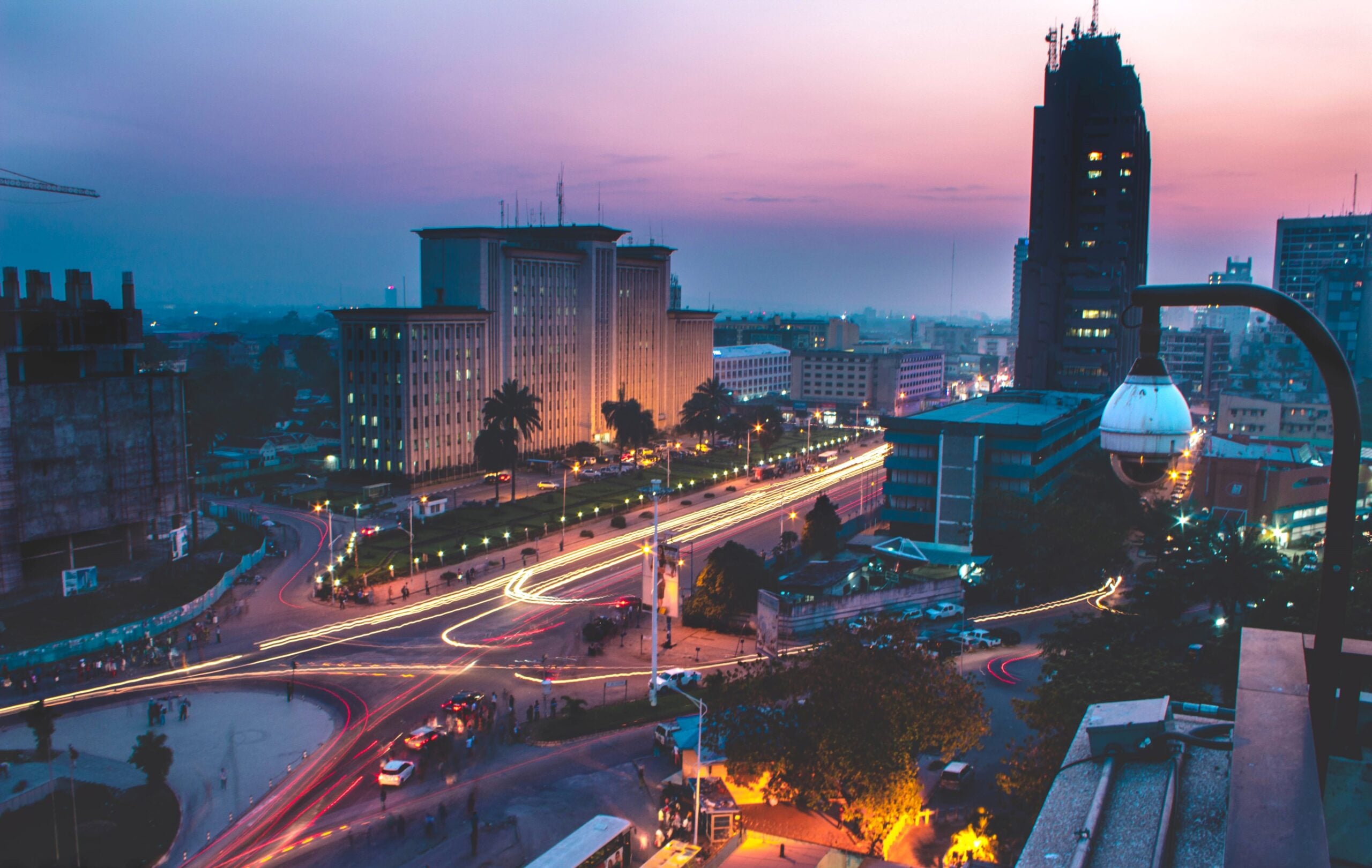 Cityscape at dusk with tall buildings, illuminated streets, and light trails from moving vehicles in one of the Biggest Cities In The World; a surveillance camera is visible in the foreground.