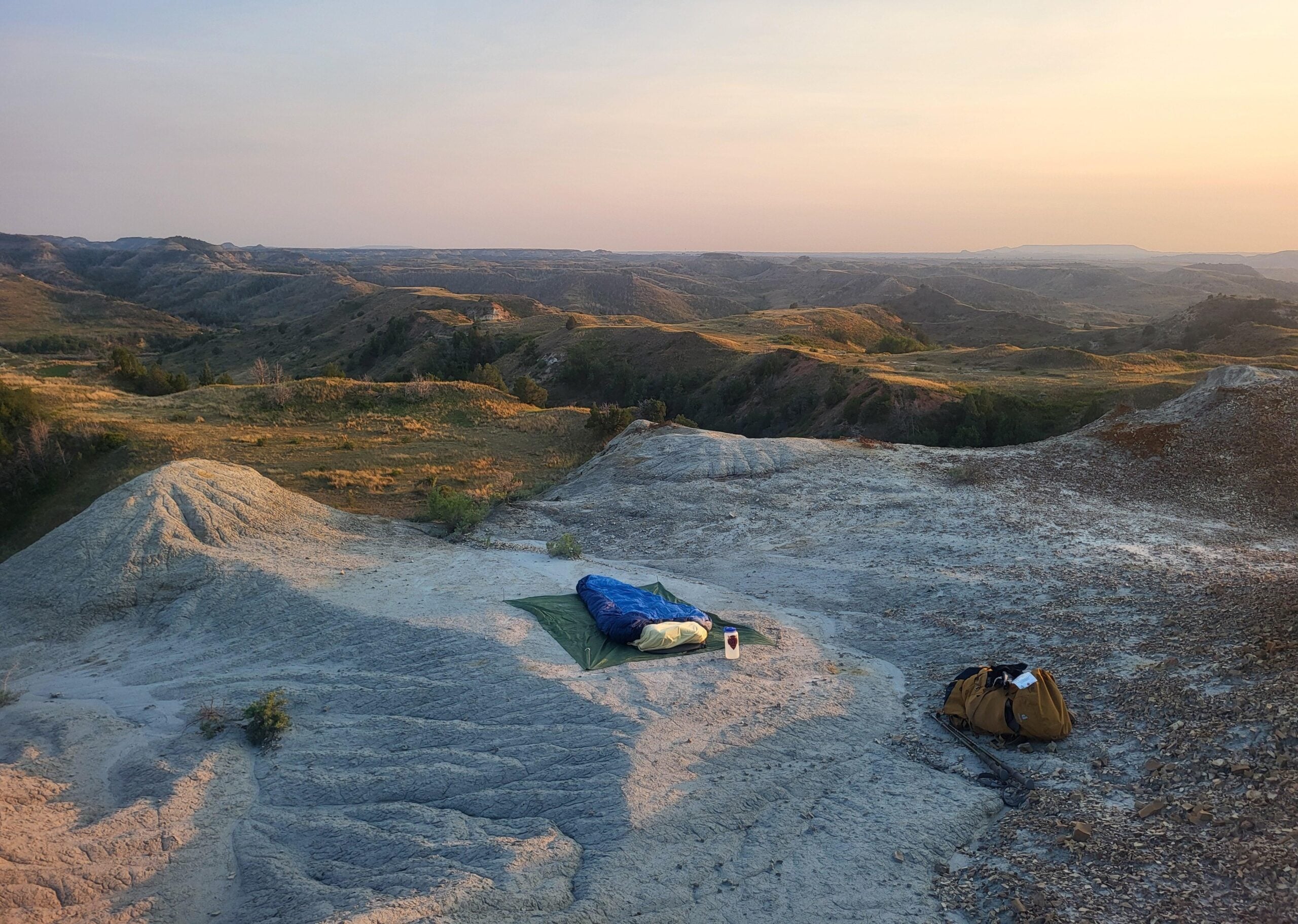 A sleeping bag, pad, cup, and backpack are set on rocky terrain overlooking rolling hills at sunrise or sunset under a clear sky.