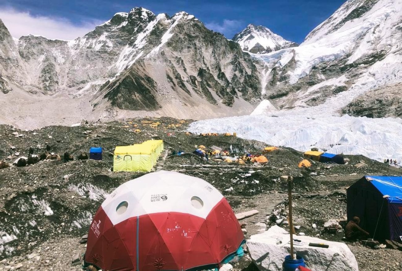 Colorful tents dot a rocky, mountainous base camp with snowy peaks in the background under a clear blue sky.