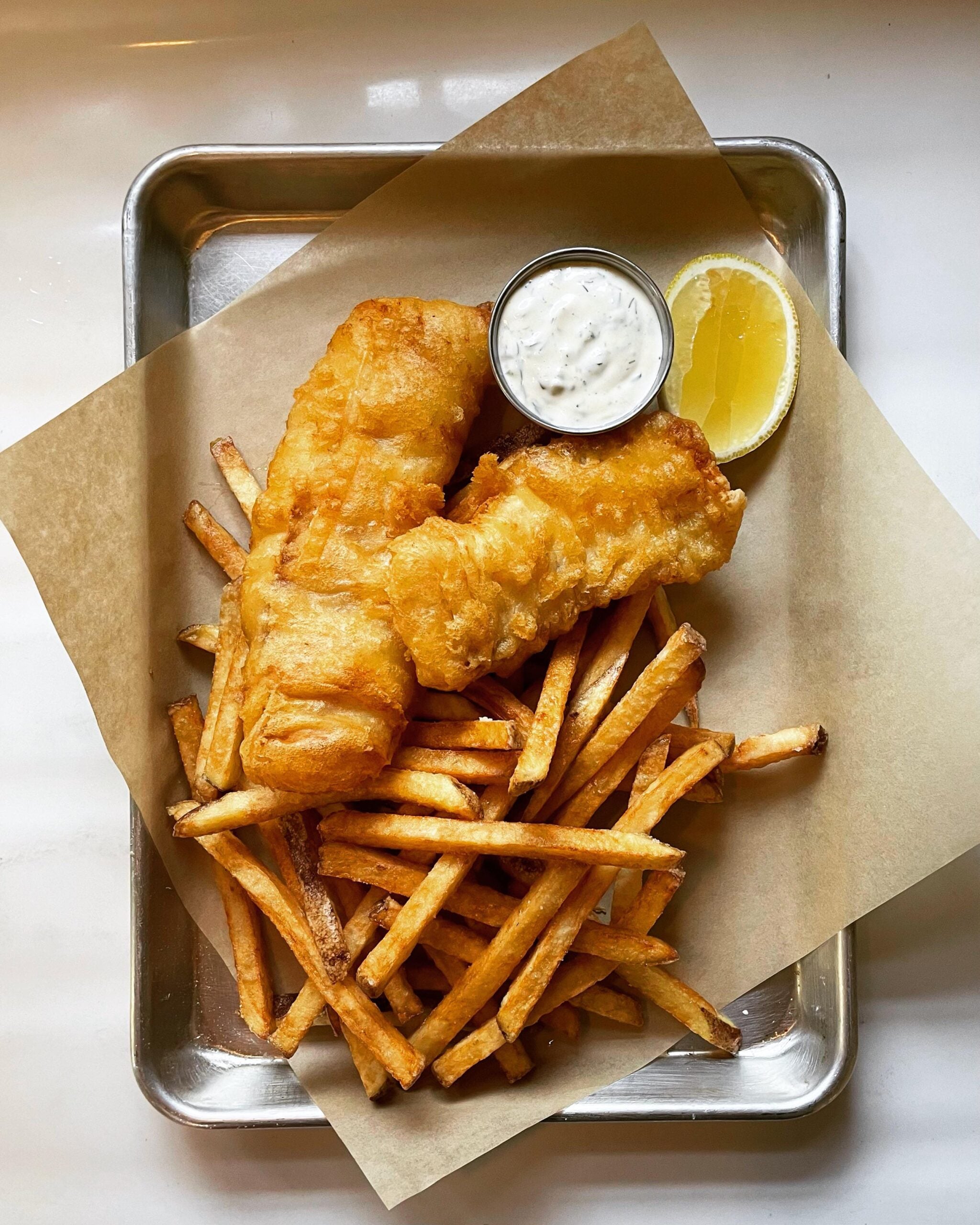 Golden fried fish fillets and French fries on parchment paper, served with a lemon wedge and a small cup of tartar sauce, all on a metal tray.