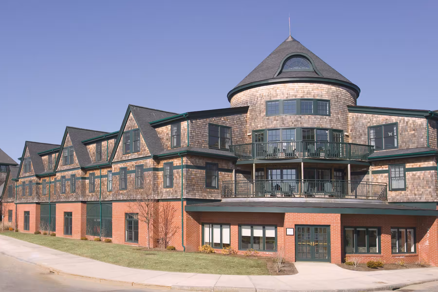 A large building with red brick on the lower level and wood shingles above, featuring green trim, multiple windows, and a distinctive round turret under a clear blue sky, evokes the timeless charm often found in classic resorts.