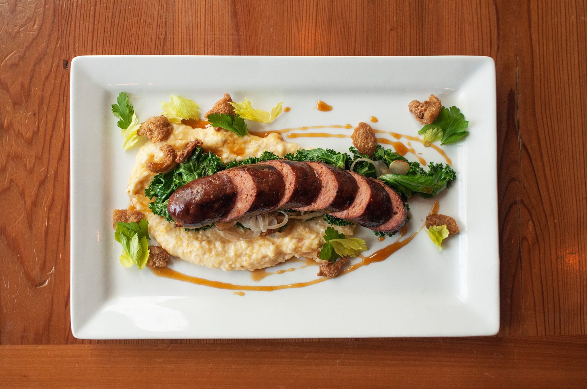 Sliced sausage on a bed of creamy polenta, sautéed greens, onions, and garnished with fresh herbs and crispy croutons, served on a rectangular white plate.