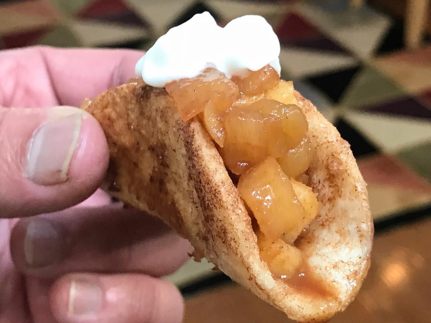 A hand holds a small taco-shaped dessert filled with diced caramelized apples and topped with a dollop of whipped cream. Geometric patterned floor is visible in the background.