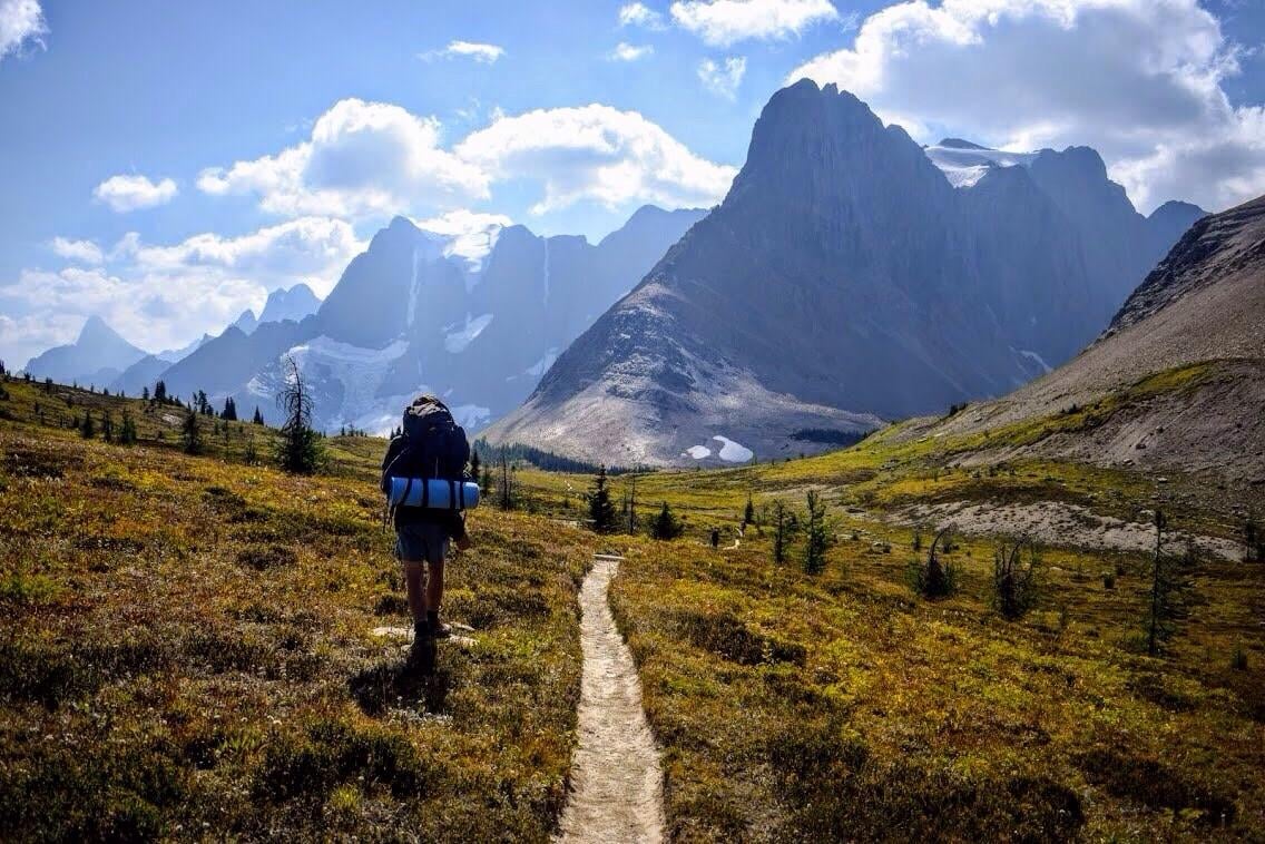 A hiker with a backpack and sleeping mat walks along a dirt trail through a mountain valley, surrounded by grassy terrain and tall, rugged peaks under a partly cloudy sky.