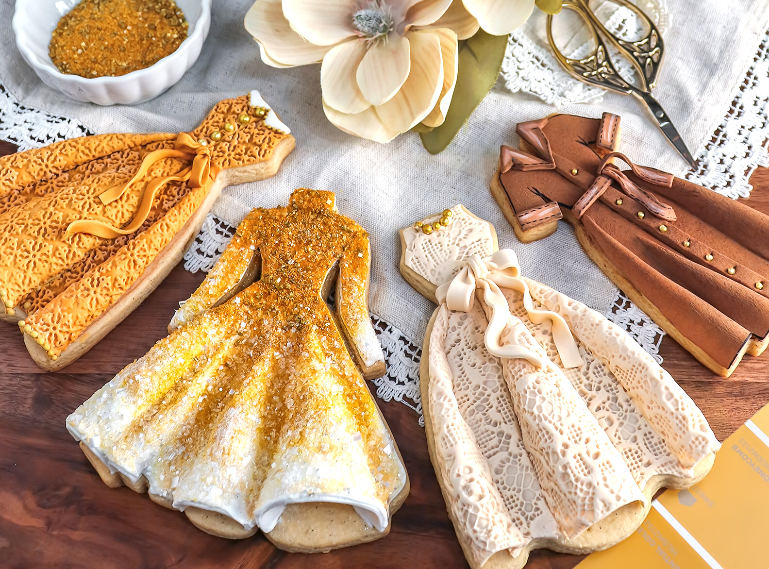 Four dress-shaped cookies decorated with gold, cream, and brown icing and glitter, arranged on a wooden table with lace, a large silk flower, scissors, and fabric swatches in the background.