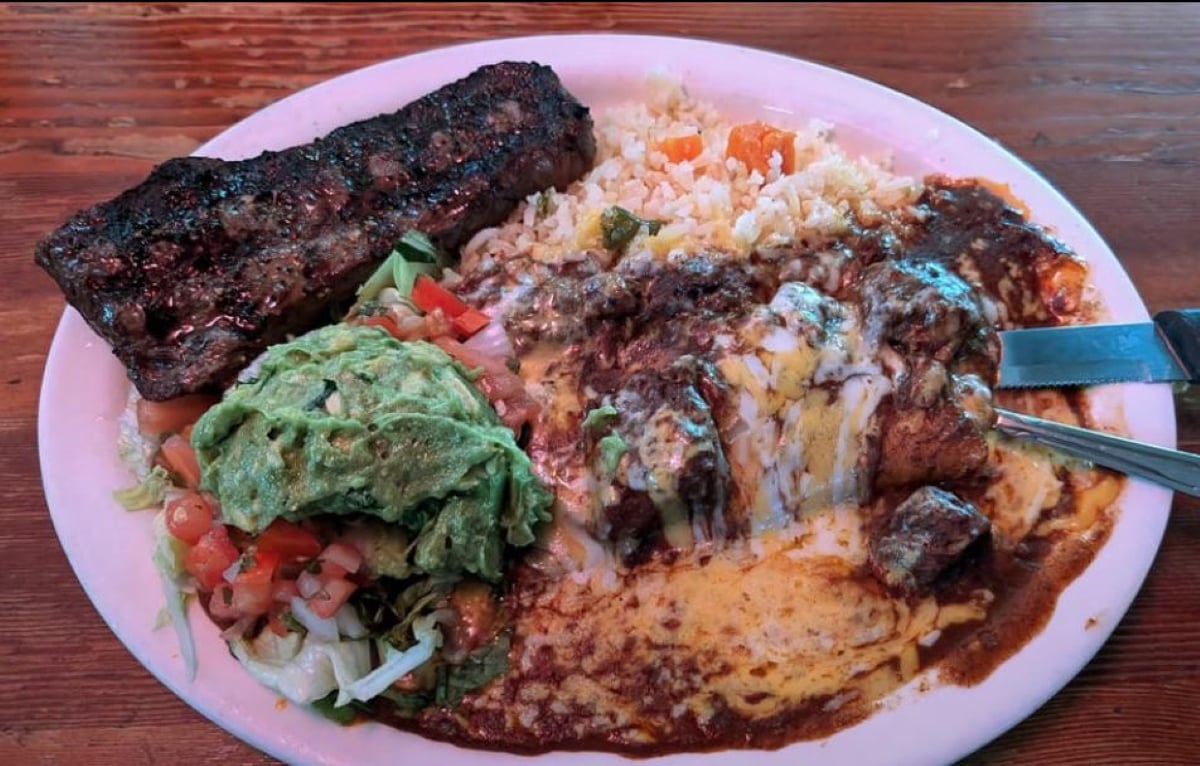 A plate with grilled steak, Mexican rice, a cheesy chili-covered enchilada, guacamole, and a fresh salad with diced tomatoes, lettuce, and onions on a wooden table.