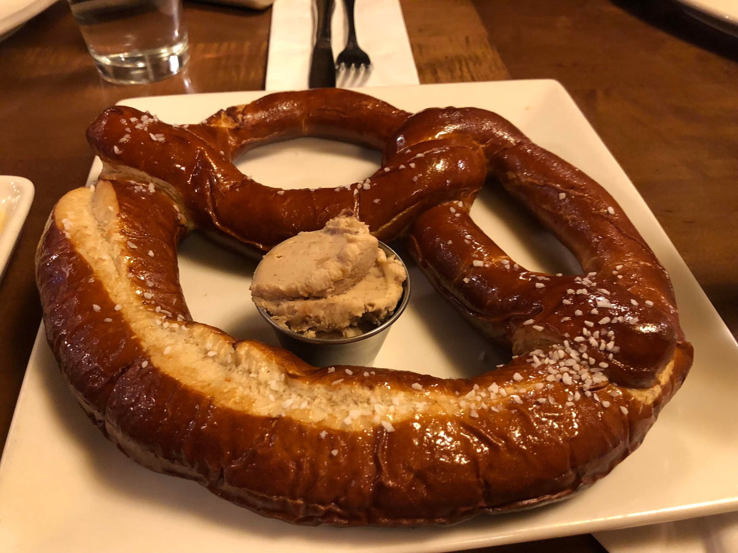 A large soft pretzel with coarse salt sits on a white plate, with a small cup of light brown dipping spread in the center. A glass of water and utensils are in the background.