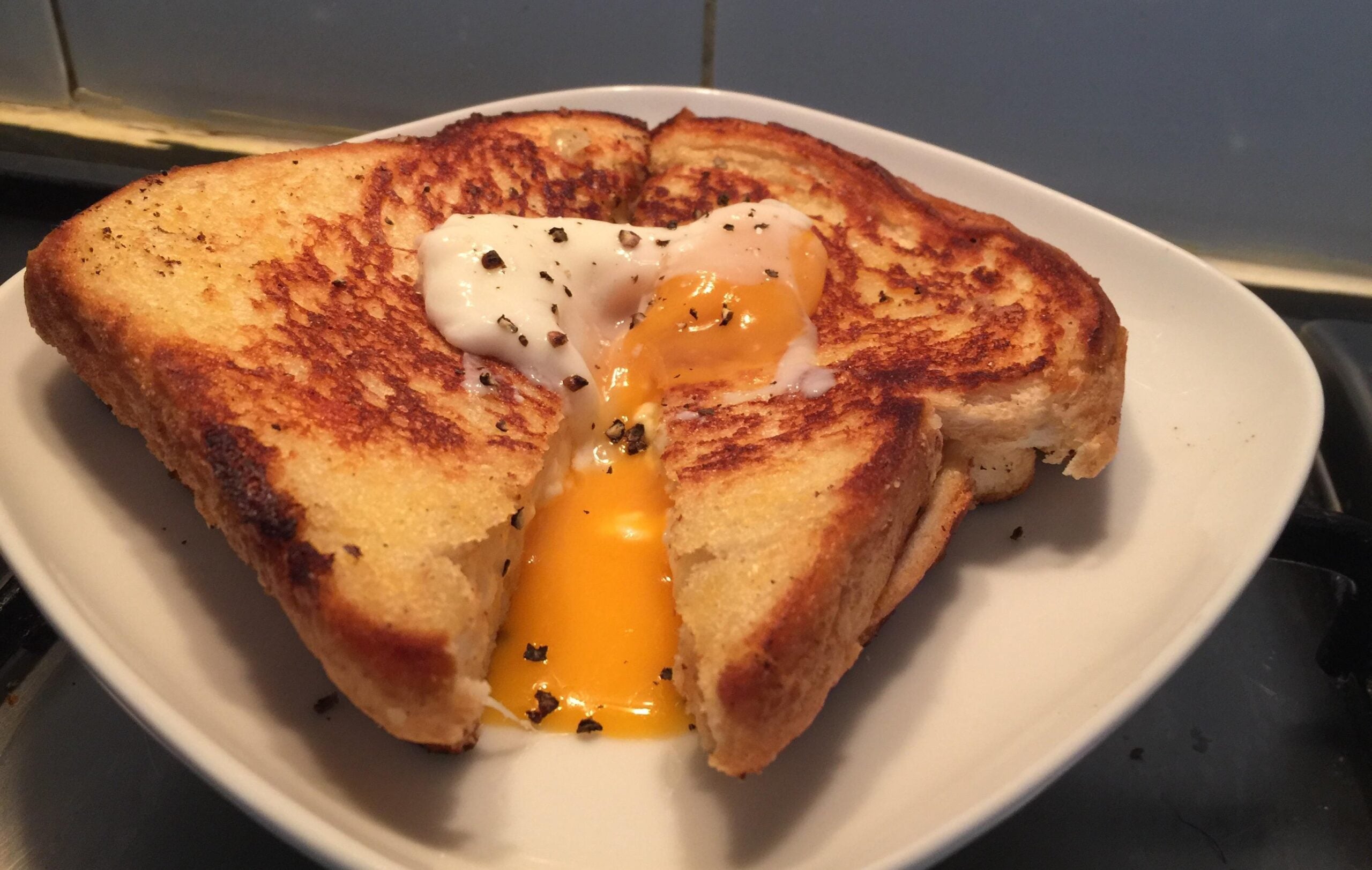 A grilled cheese sandwich on a white plate is cut open, revealing a runny egg yolk and sprinkled black pepper on top—a delicious twist on classic Grilled Cheese Sandwiches.
