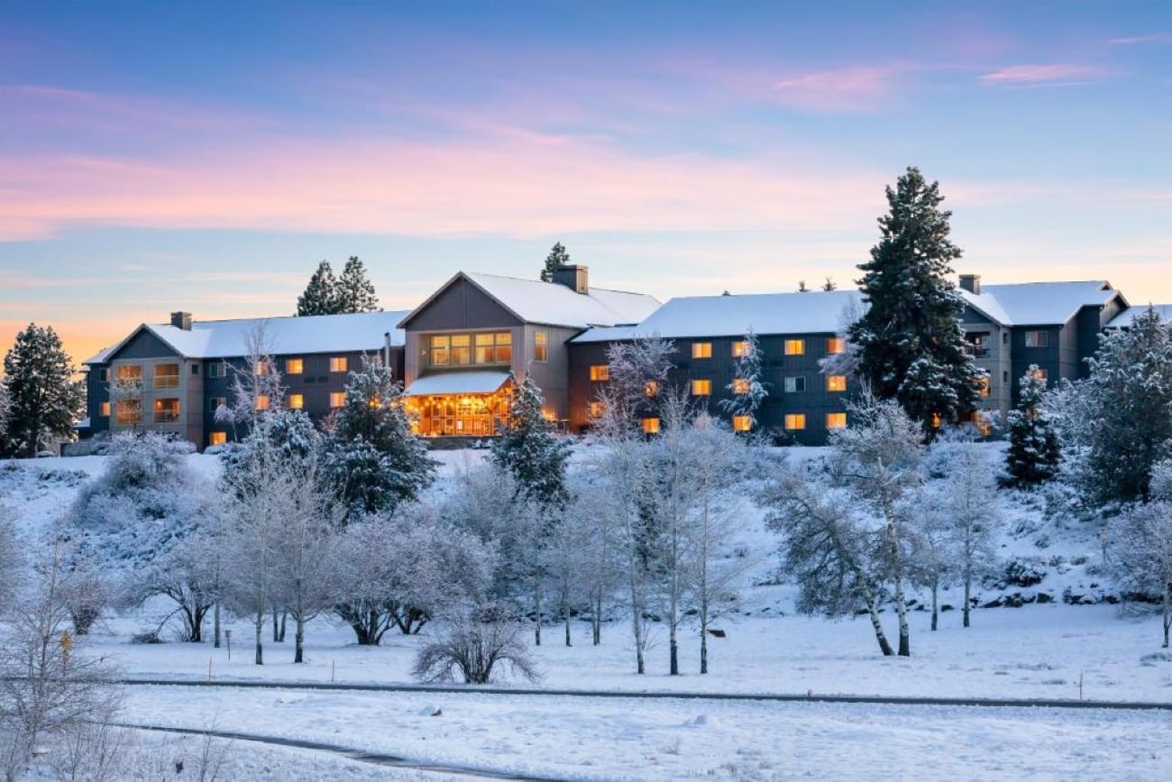 A large lodge-style building, reminiscent of upscale resorts, with warm glowing windows sits on a snowy hill surrounded by frosted trees at sunset, under a pink and blue sky.
