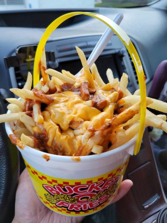 A hand holds a bucket filled with French fries covered in melted cheese and chili, with a fork inserted. The bucket has a yellow handle and a &ldquo;Ricky&rsquo;s Fast Fries&rdquo; logo. The image is taken inside a car.