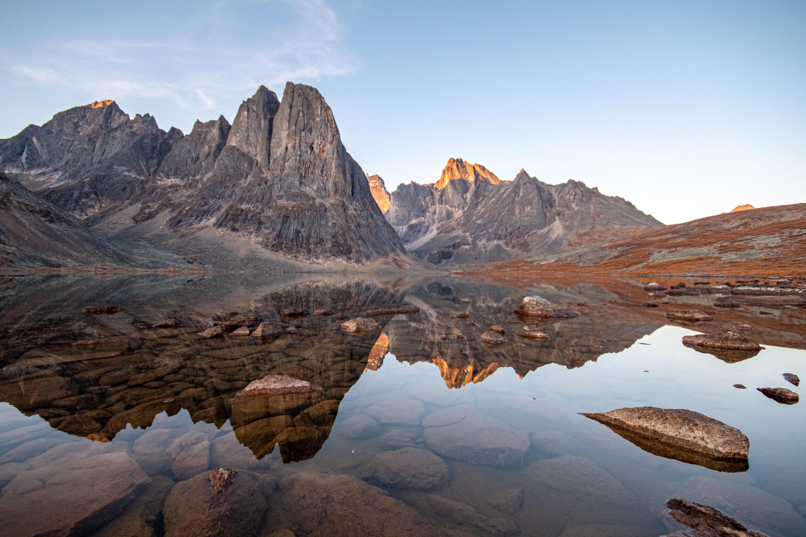 Jagged mountain peaks are reflected in a calm, clear lake with rocks visible beneath the surface, under a blue sky with soft evening or morning light.