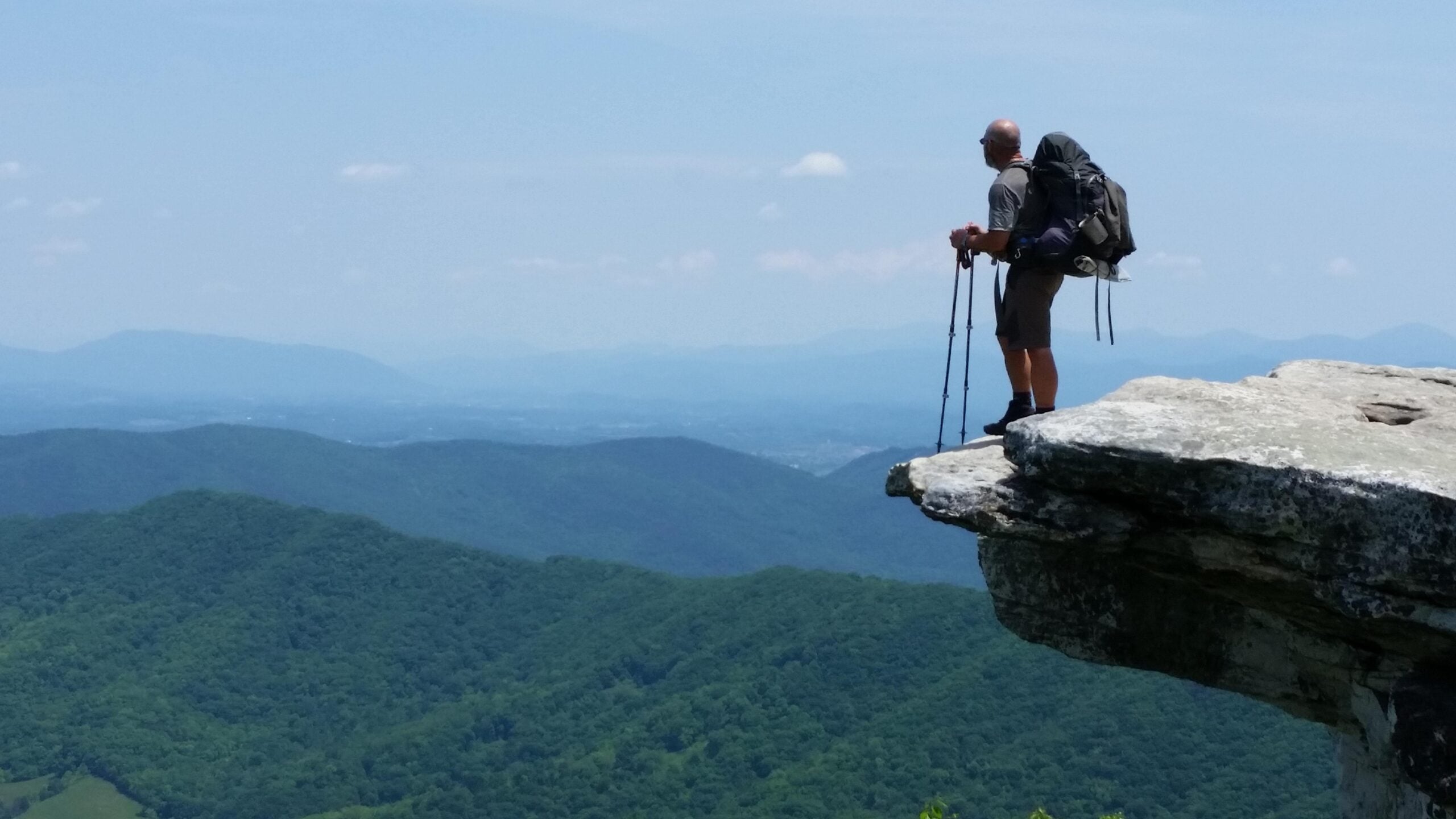 A hiker with a backpack and trekking poles stands on the edge of a rocky cliff, looking out over rolling green mountains under a blue sky, capturing the breathtaking scene for photos hikers often posted after adventurous journeys.