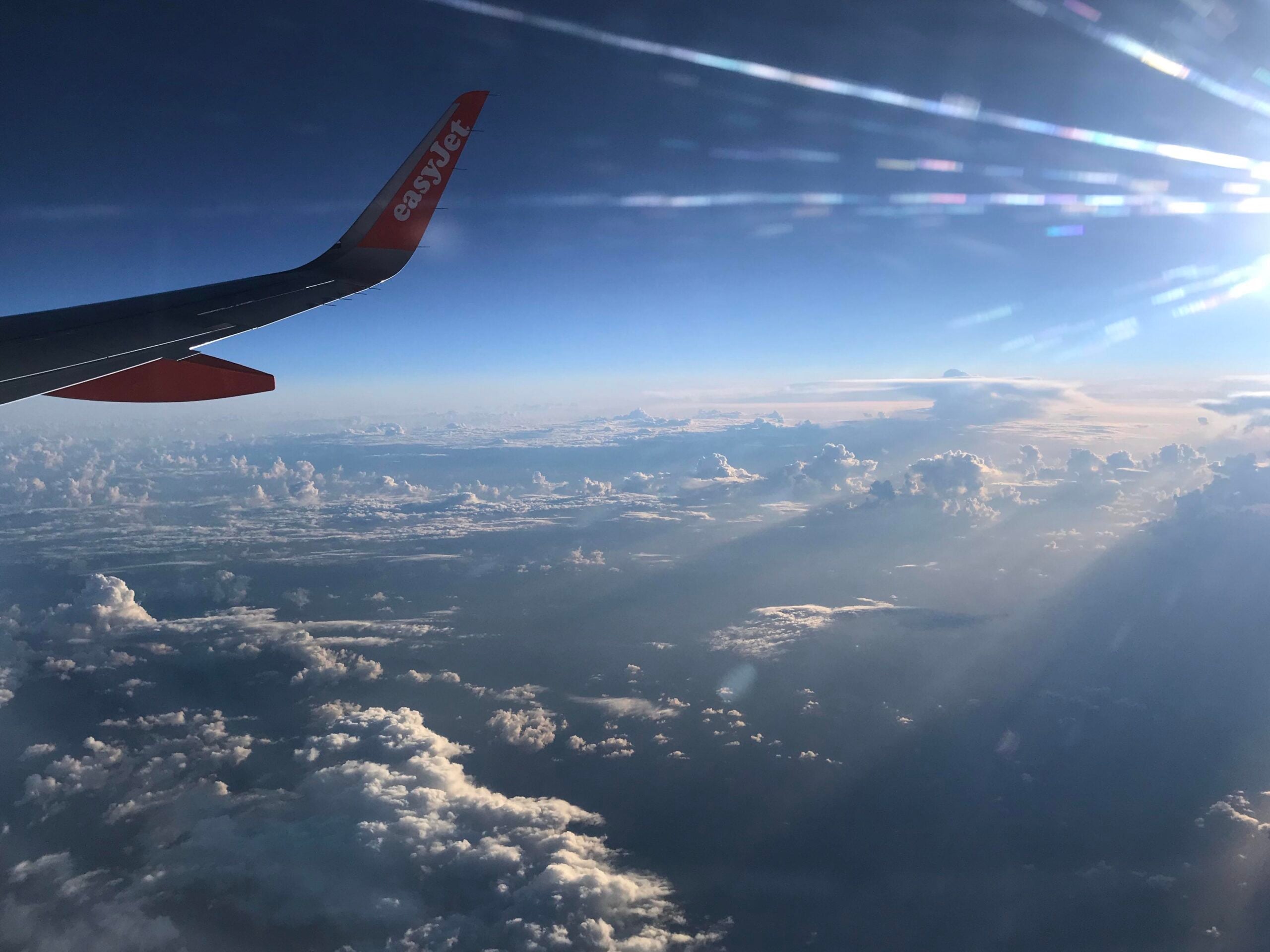 View from airplane windows showing an easyJet wing, sun rays, and clouds below against a blue sky.