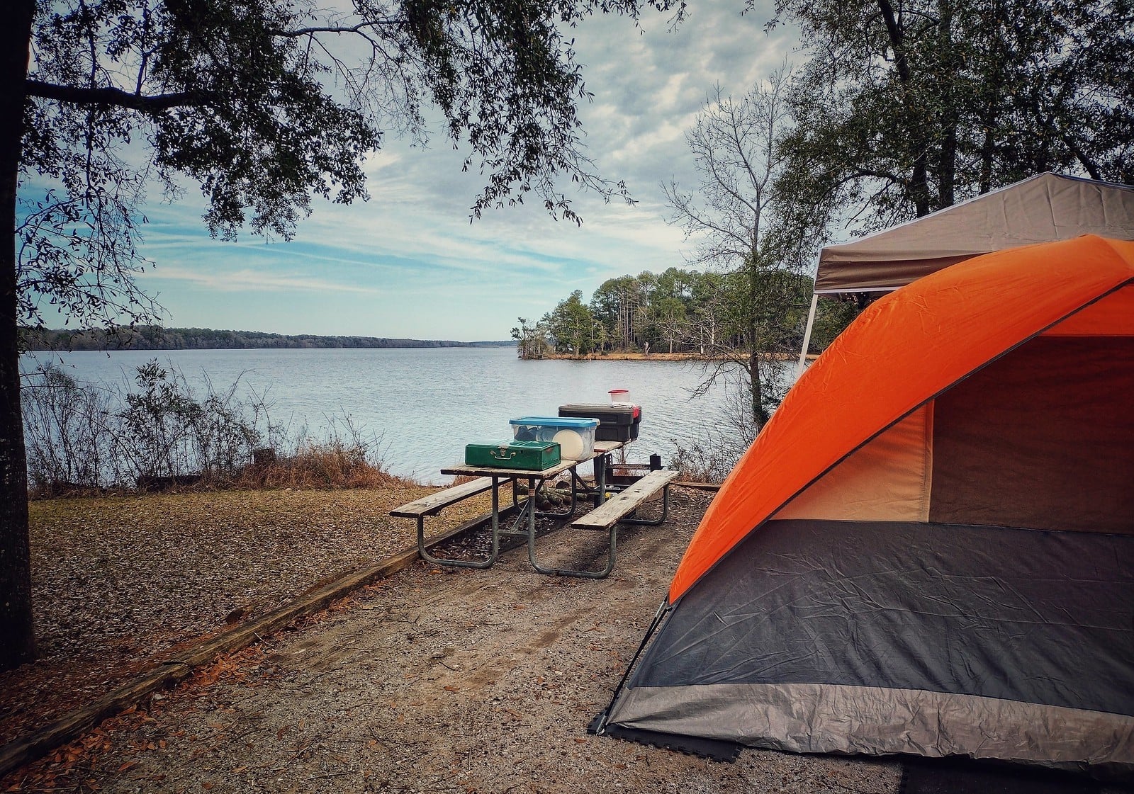 An orange and gray tent is set up by a lake. Nearby, a picnic table holds camping gear, including a green stove and cooler. Trees surround the peaceful lakeside campsite under a partly cloudy sky.