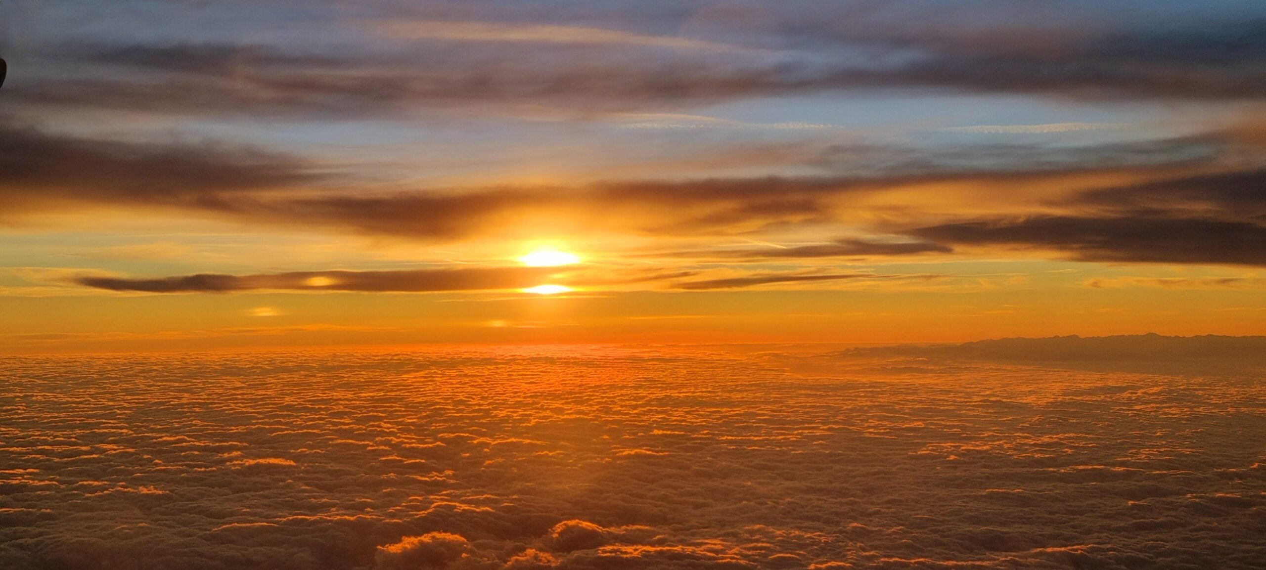 A bright orange sun sets over a dense layer of clouds, casting warm, golden light across the sky and creating dramatic hues of yellow, orange, and blue among scattered clouds—an awe-inspiring view often admired through airplane windows.