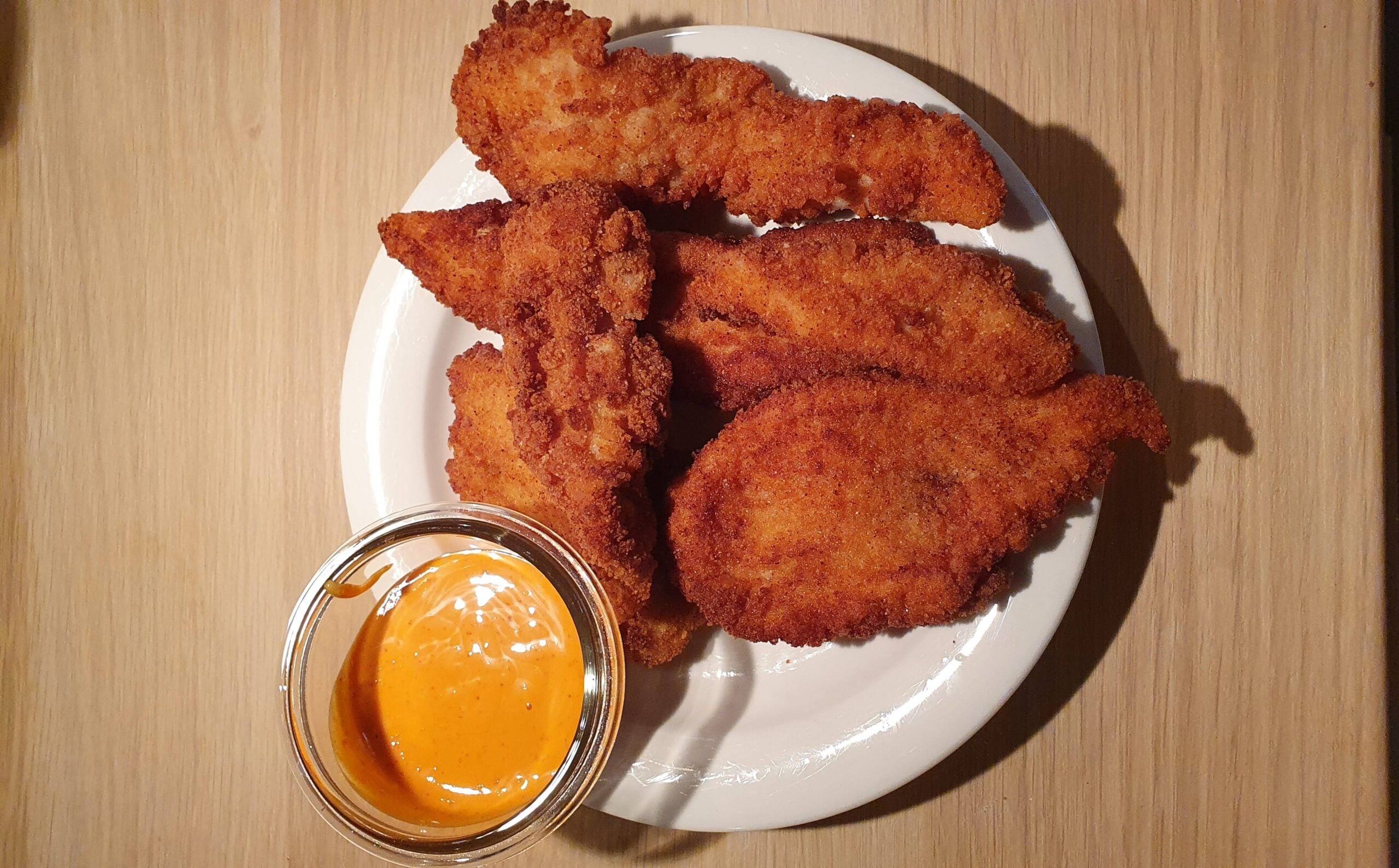 A white plate with five pieces of breaded fried chicken and a small glass bowl of orange dipping sauce on a wooden table.