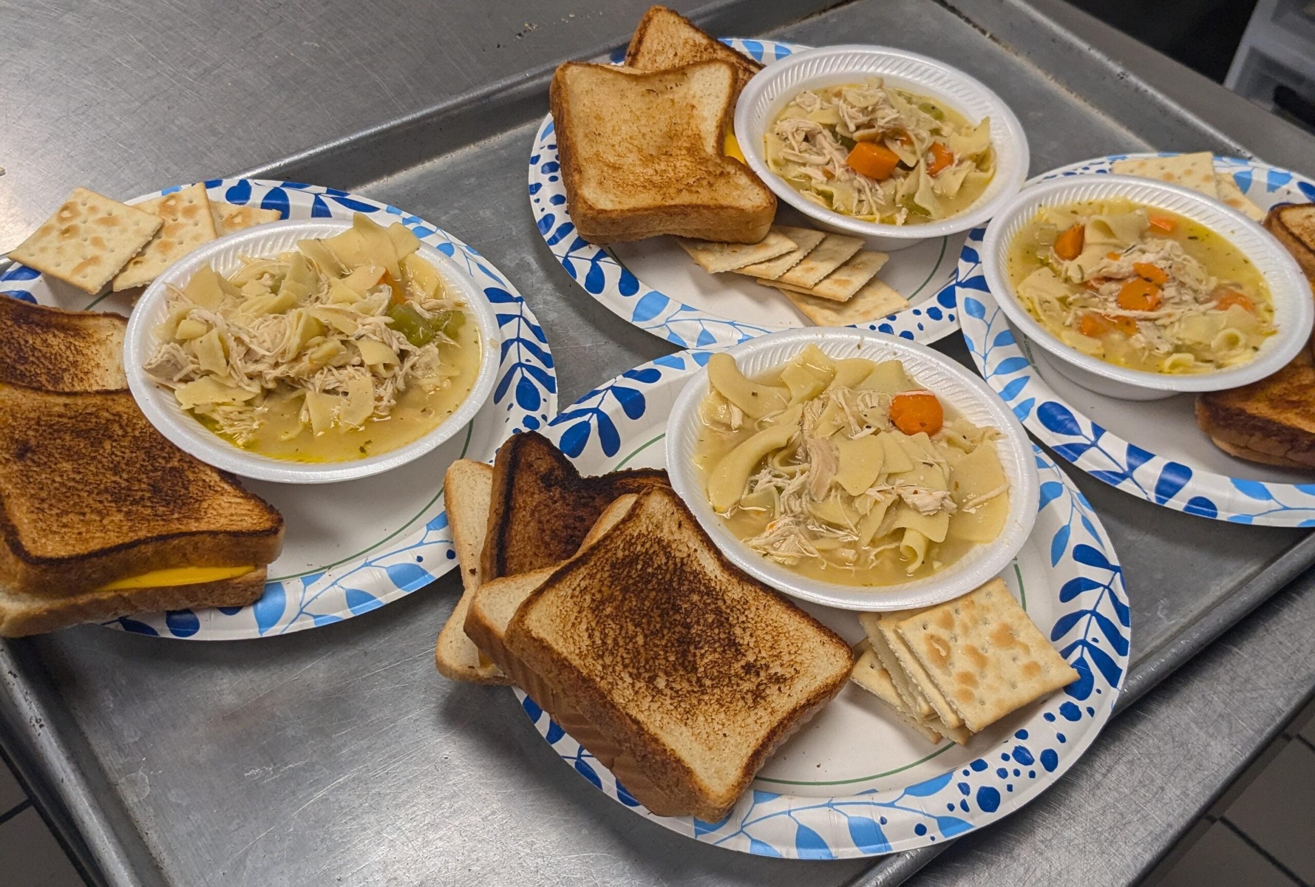 Four paper plates each hold a bowl of chicken noodle soup, square crackers, and slices of toasted bread with cheese reminiscent of classic grilled cheese sandwiches, all arranged on a metal tray.