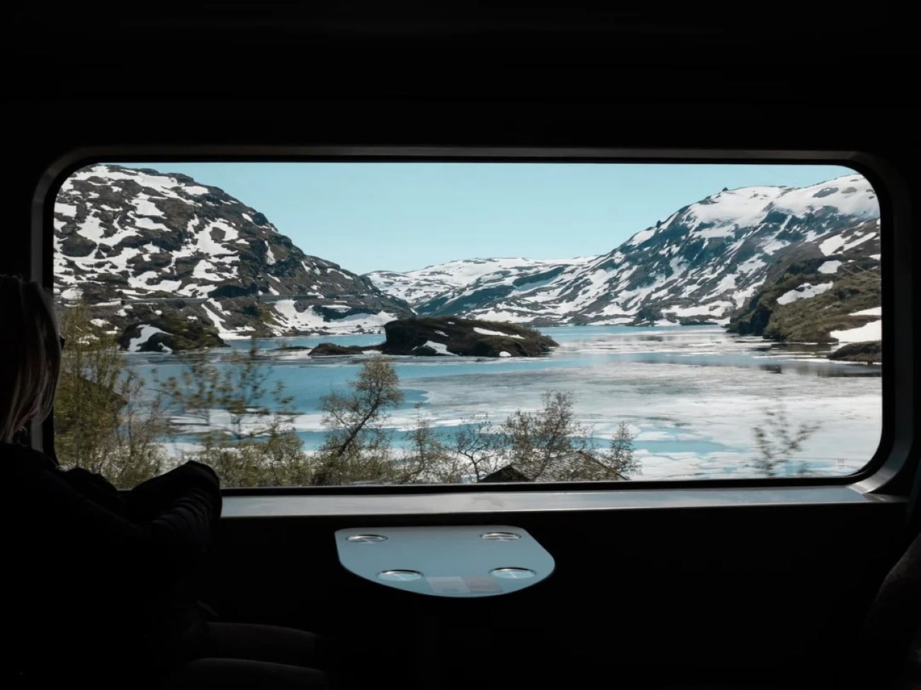 A person sits inside a train looking out a large window at snowy mountains, a partially frozen lake, and clear blue sky.