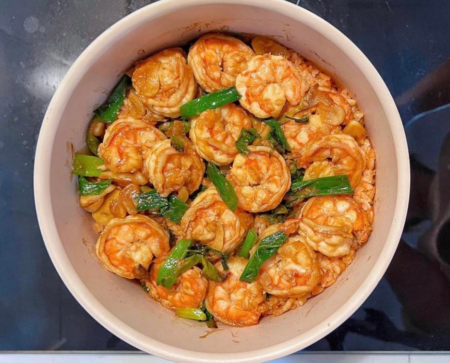 A pot filled with cooked shrimp, green onions, and sliced garlic in a reddish sauce, sitting on a stovetop.