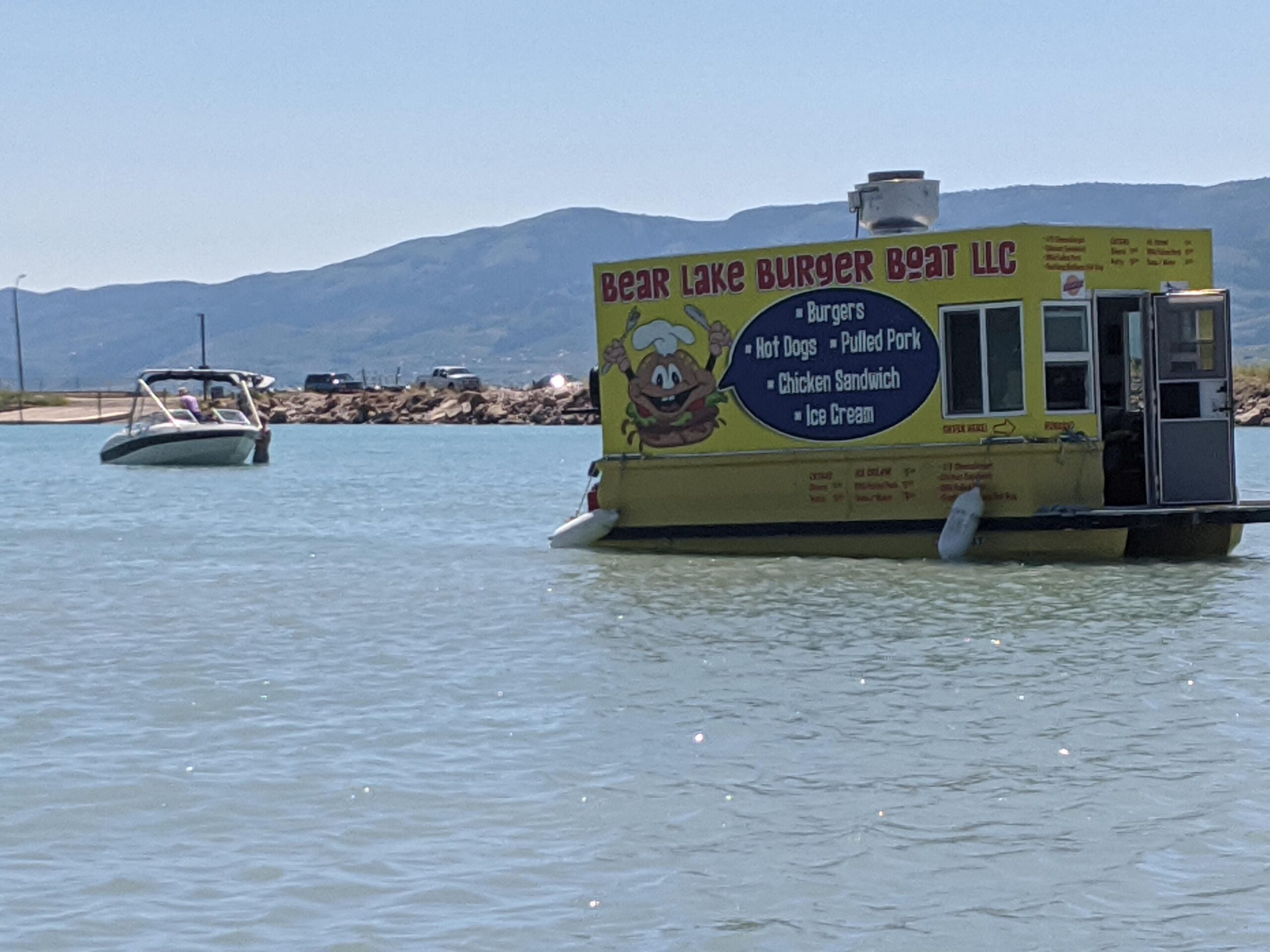 A yellow floating food stand labeled “Bear Lake Burger Boat LLC” is on a lake, with a speedboat and mountains visible in the background under a clear sky.