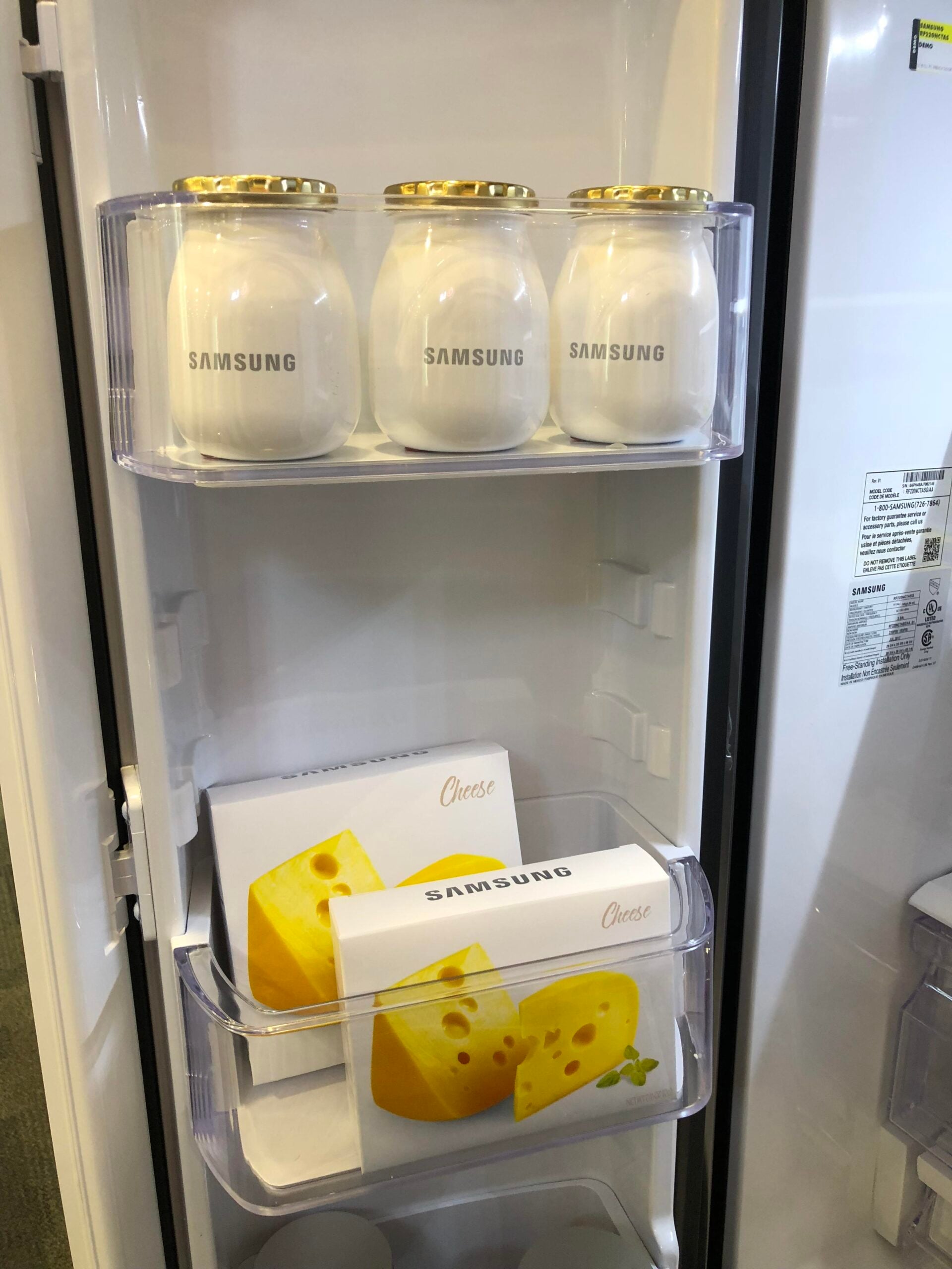 Three jars labeled “Samsung” on the top fridge shelf; below, two boxes with images of cheese, also labeled “Samsung,” are on the second shelf inside the refrigerator door.