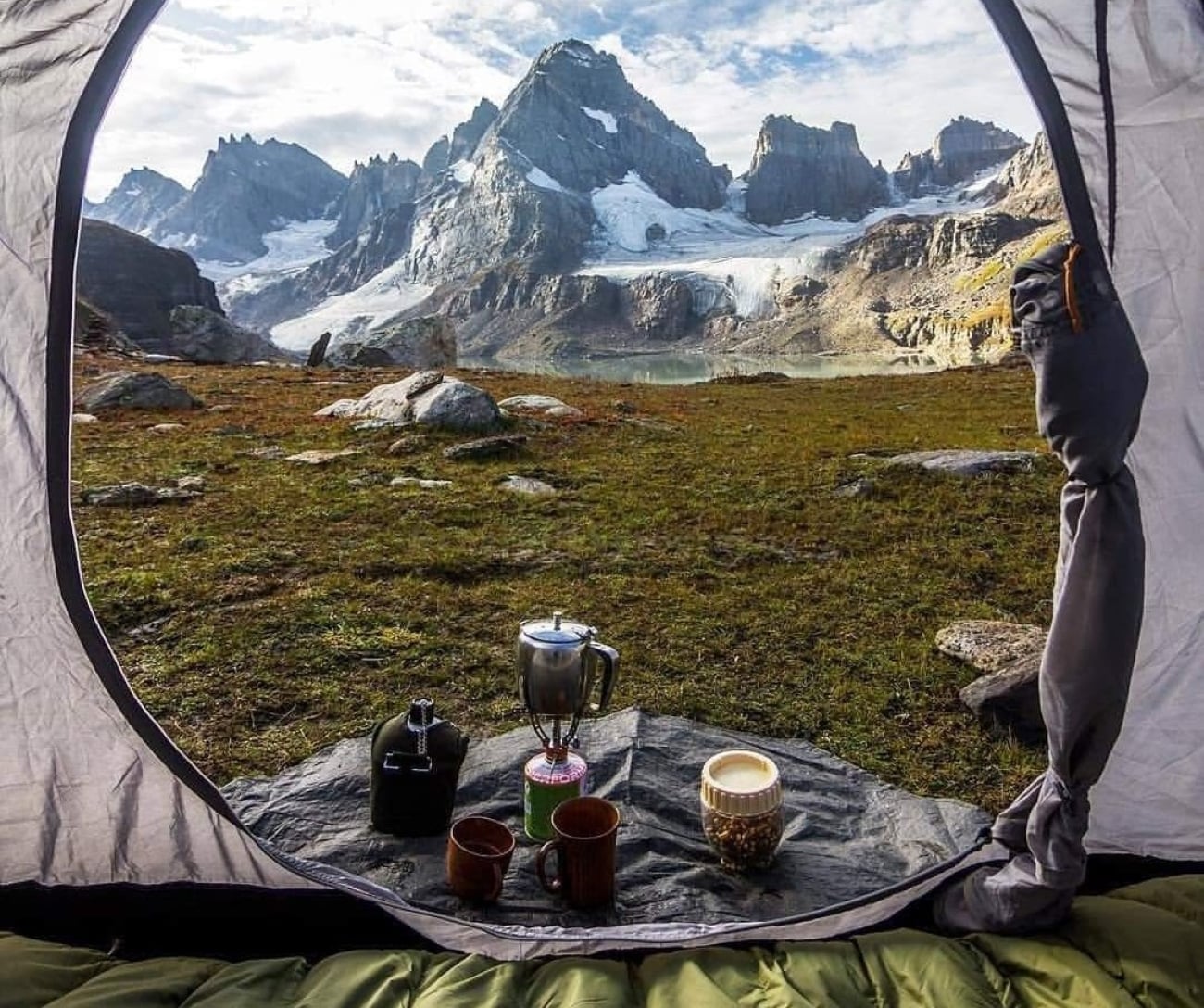 View from inside a tent showing coffee cups, a French press, and a thermos on a rock, with majestic snow-capped mountains and grassy terrain outside under a partly cloudy sky—just like the stunning photos hikers posted after their latest adventures.