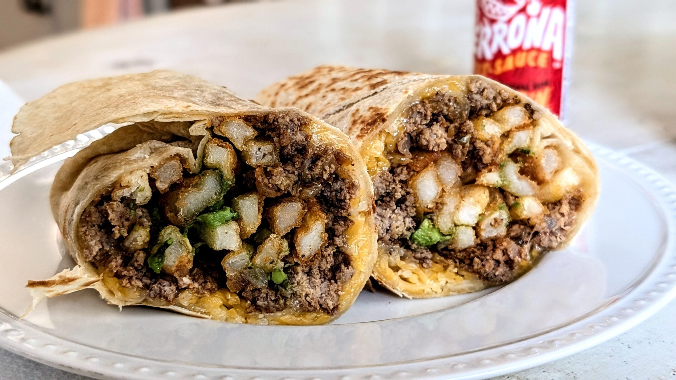 A burrito cut in half, showing ground beef, French fries, cheese, and green peppers inside, on a white plate with a red hot sauce bottle in the background.
