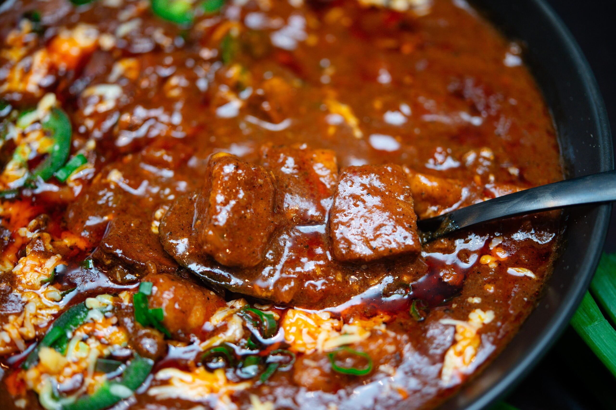 A close-up of a spoonful of thick, rich chili with chunks of meat, garnished with sliced green jalapeños and melted cheese in a black bowl.