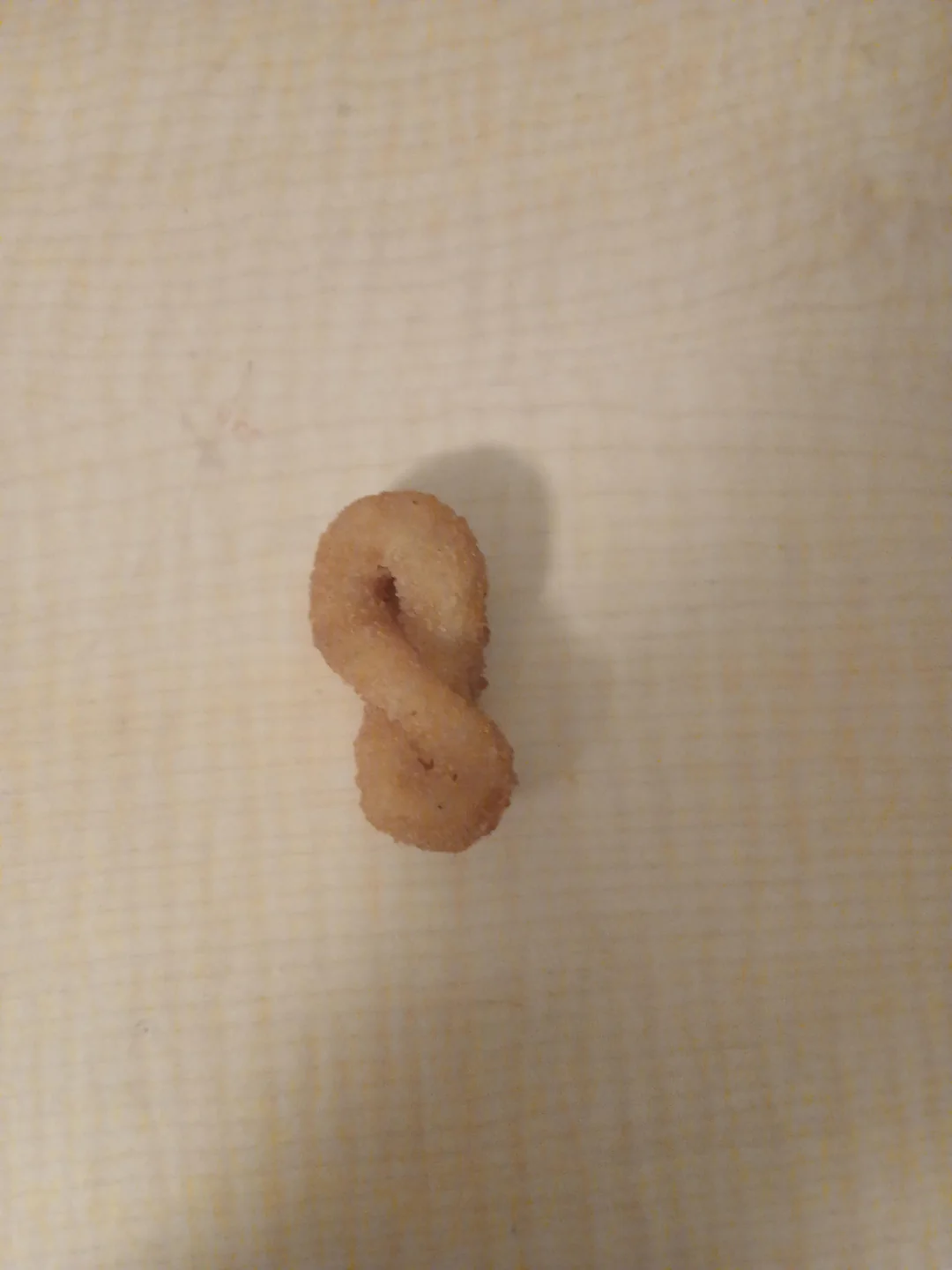 A single, small, fried snack shaped like the number eight or an infinity symbol sits on a light-colored, textured surface.