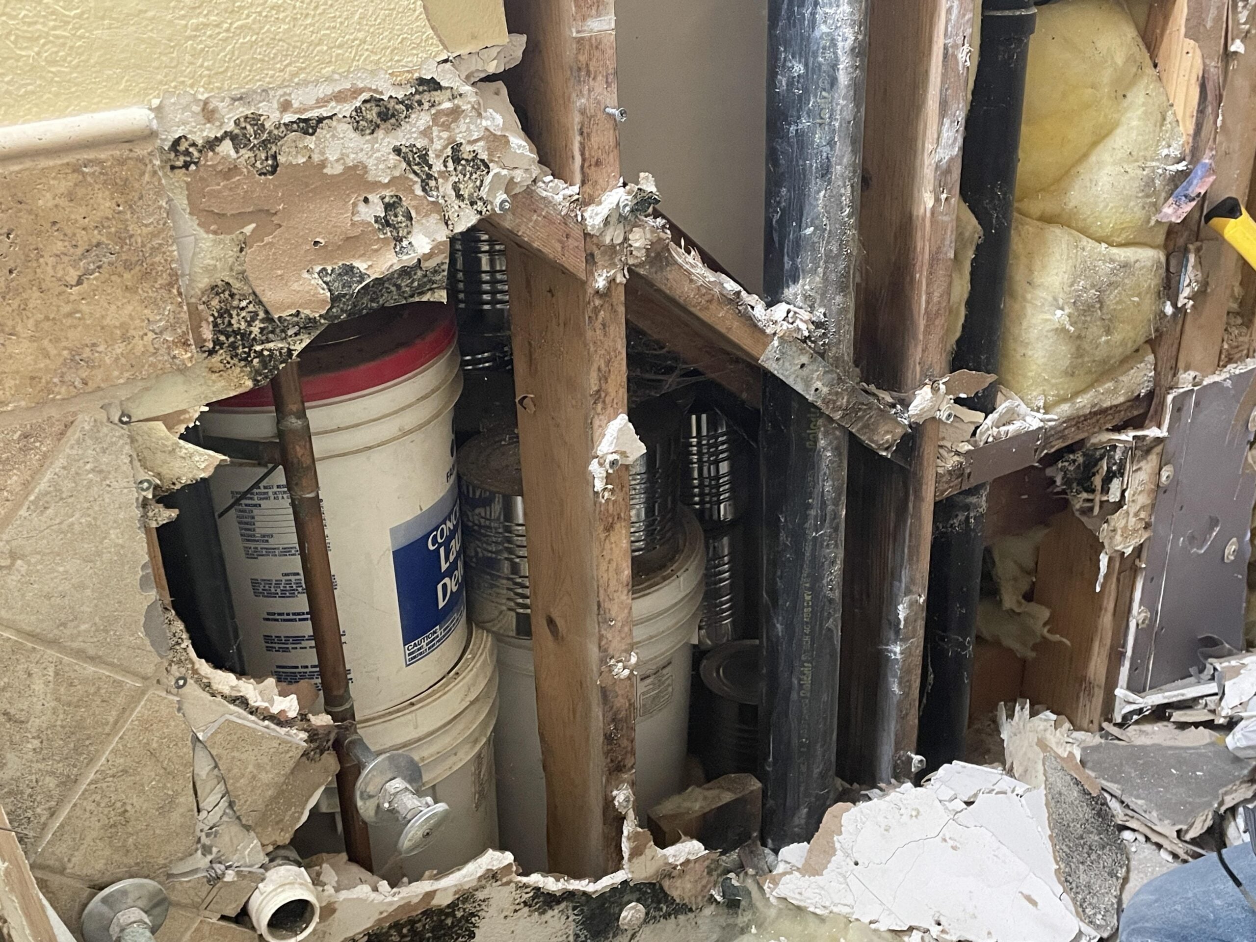 A wall with drywall and tiles removed, exposing wooden studs, pipes, insulation, and large white buckets inside a damaged area, suggesting repair work or water damage.