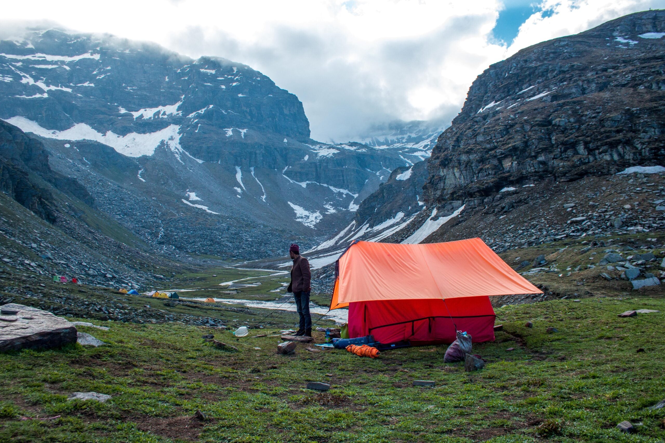 A person stands near an orange tent pitched on green grass, surrounded by rocky mountains with patches of snow under a cloudy sky. Camping gear is scattered nearby.