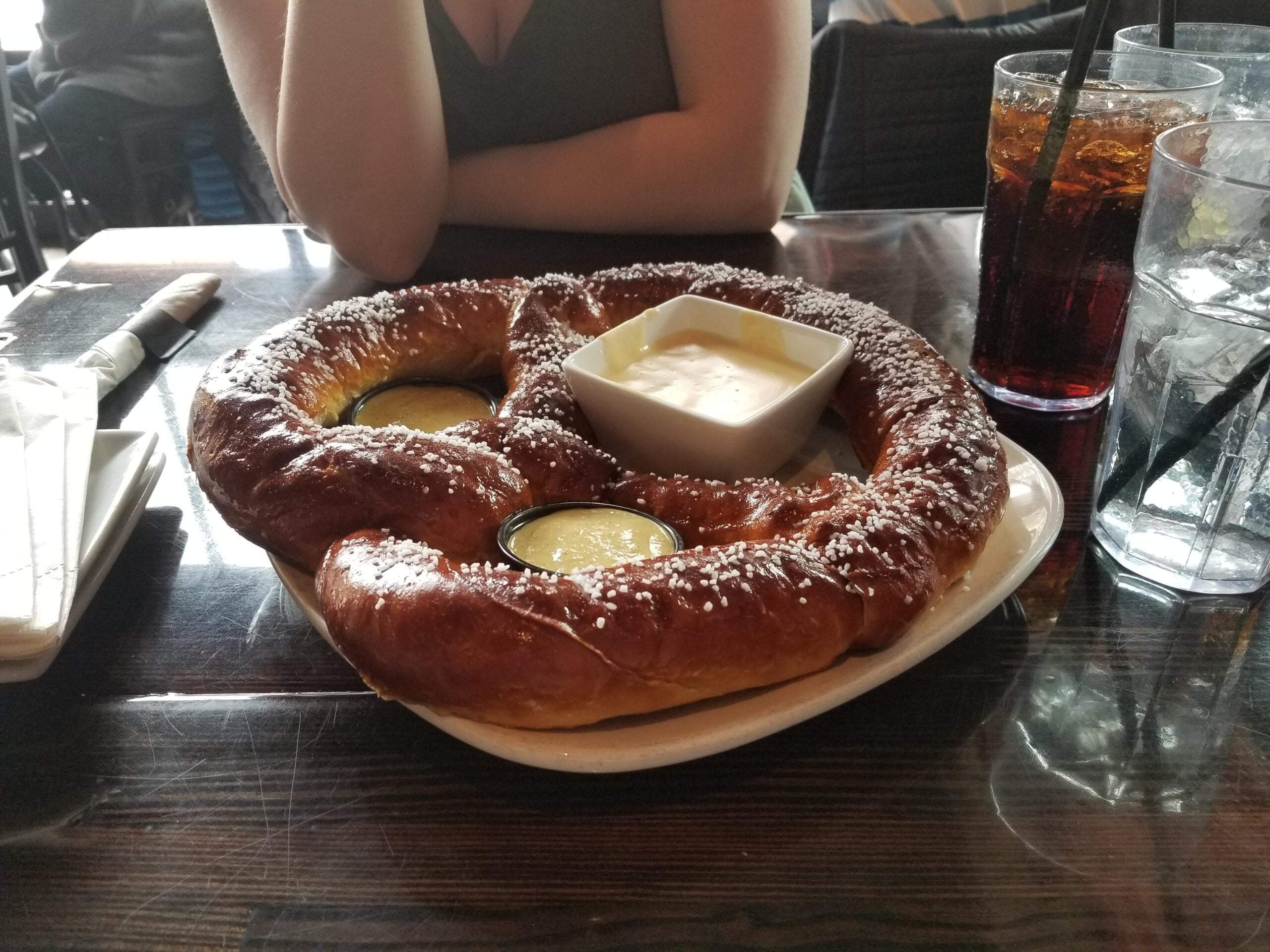 A large salted pretzel on a plate with two dipping sauces, one in a square bowl, sits on a dark wooden table. A person is seated behind the plate, with drinks and napkins nearby.