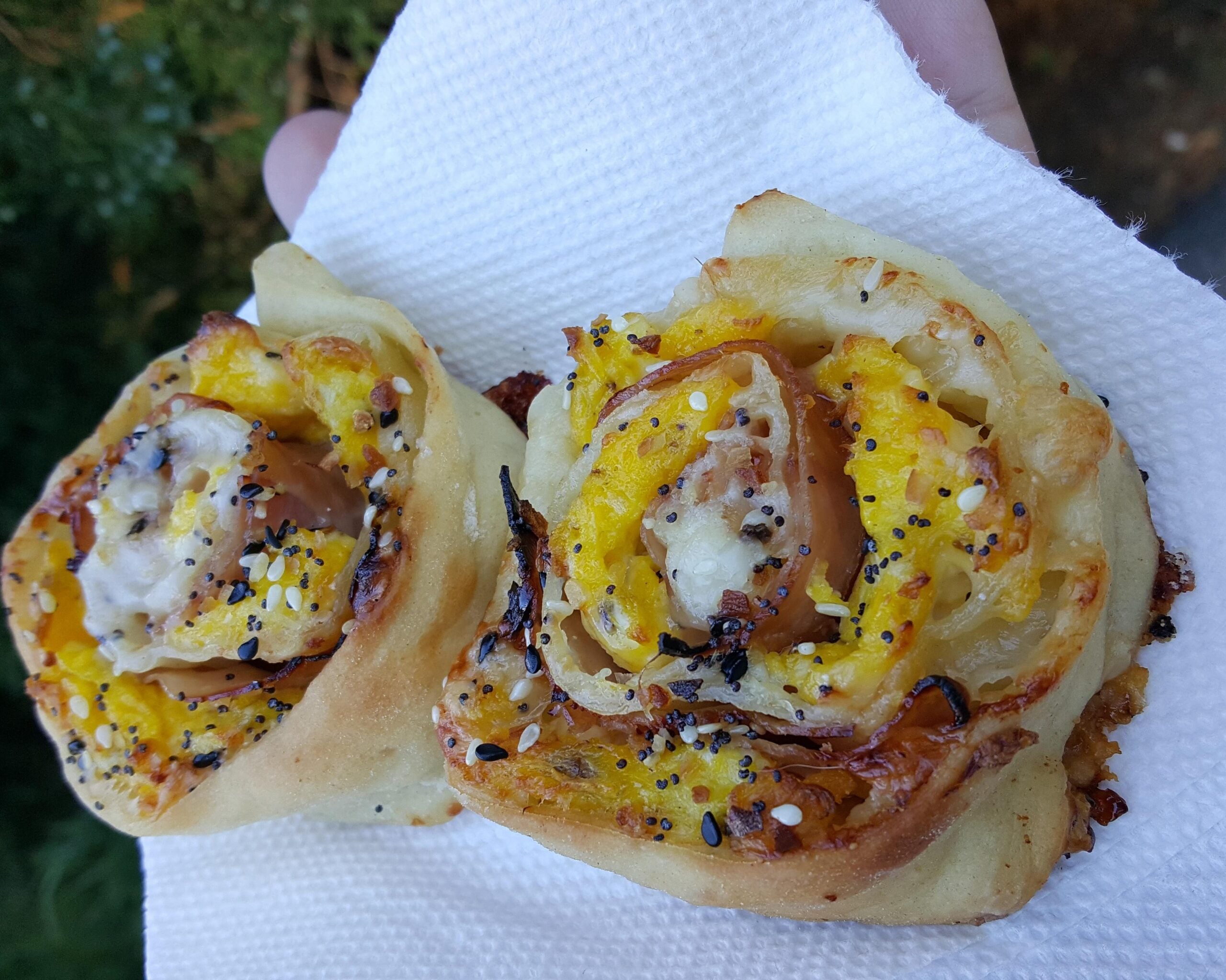 Two savory pastry rolls with cheese, ham, and seasoning, resting on a white paper towel, held outdoors with greenery blurred in the background.