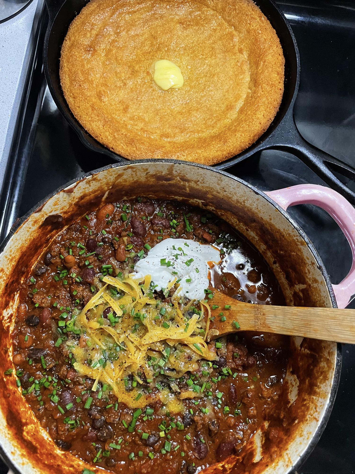 A pot of chili topped with shredded cheese and chives sits next to a skillet of golden cornbread with a pat of butter melting in the center. A wooden spoon rests in one of these chili recipes.
