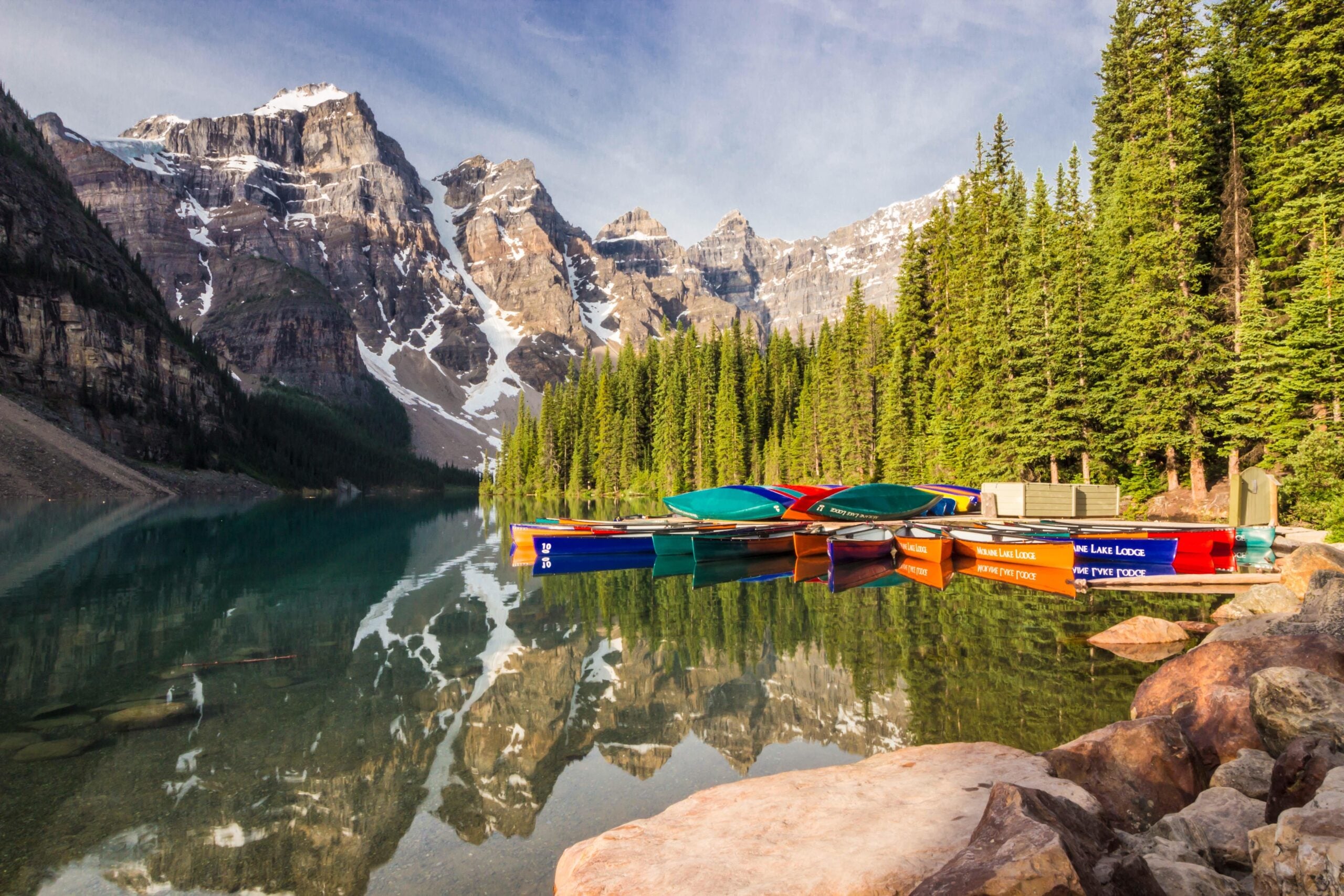 Canoes docked on a clear, reflective lake surrounded by pine trees and snow-capped mountains under a blue sky.