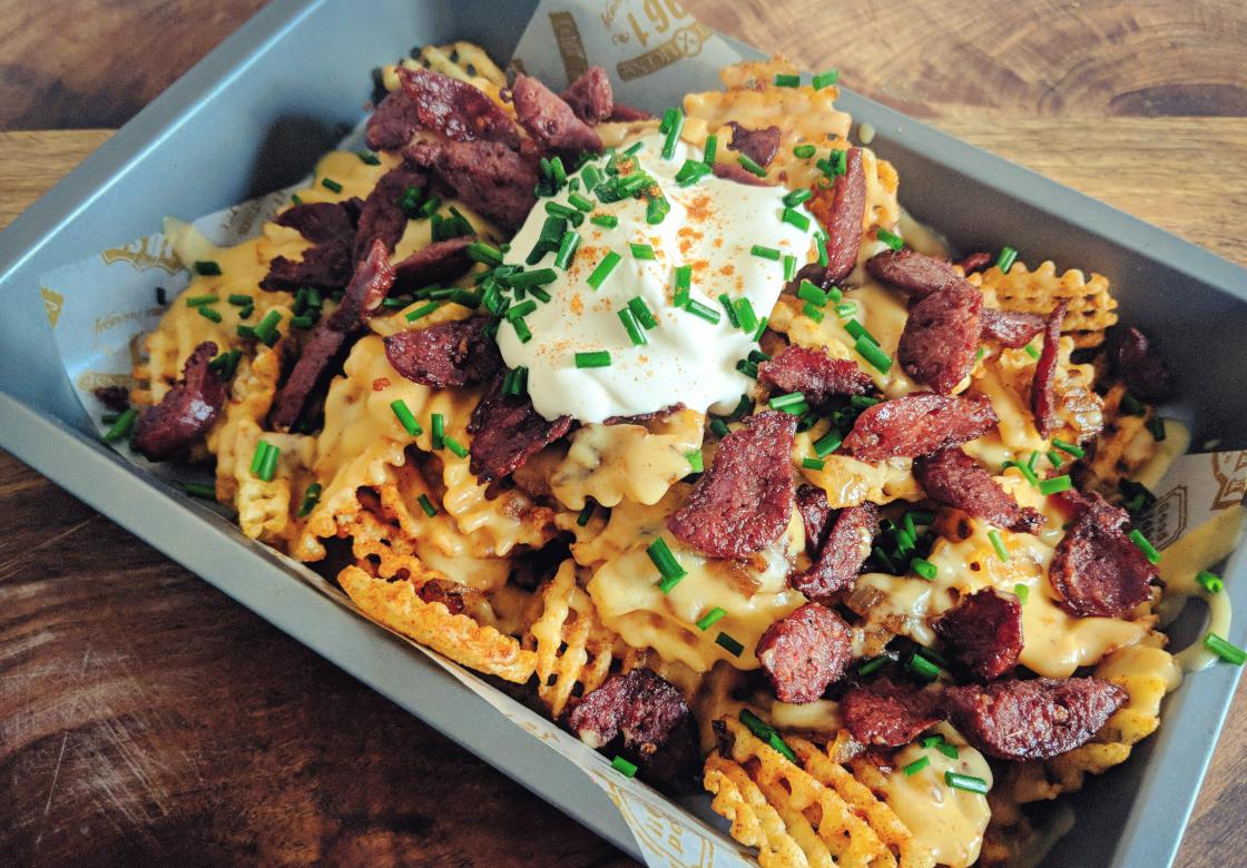 Waffle fries topped with melted cheese, sliced sausage, a dollop of sour cream, and chopped chives, served in a gray tray on a wooden surface.