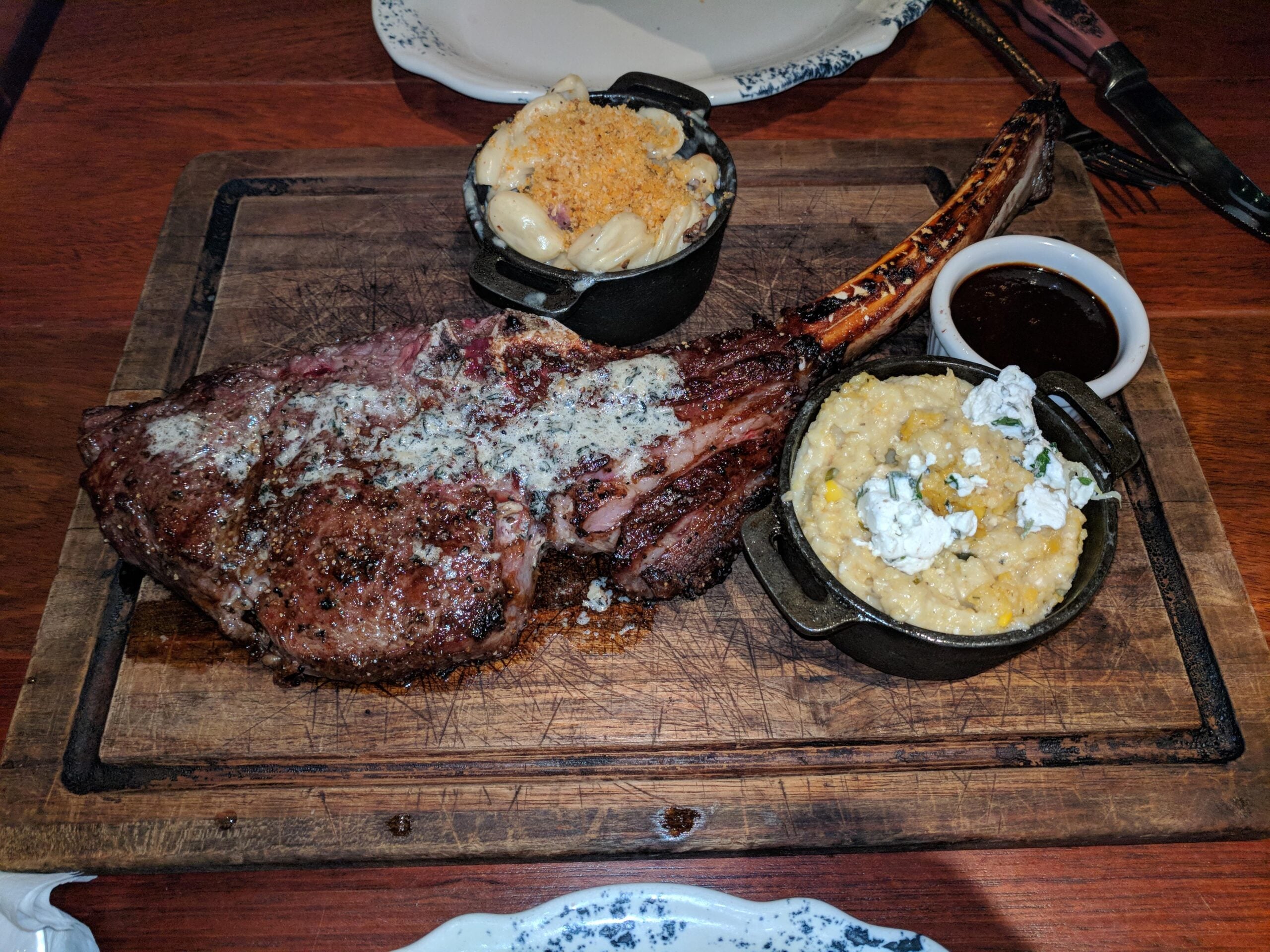 A large grilled tomahawk steak on a wooden board, served with small bowls of macaroni and cheese, creamy grits topped with cheese, and a side of brown sauce.